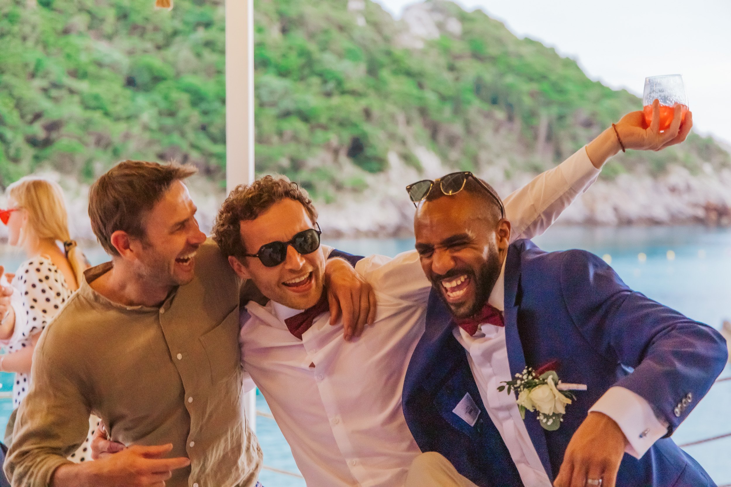 Three men celebrating and having fun, two of them wearing suits and bow ties, outdoors near water and green hills.