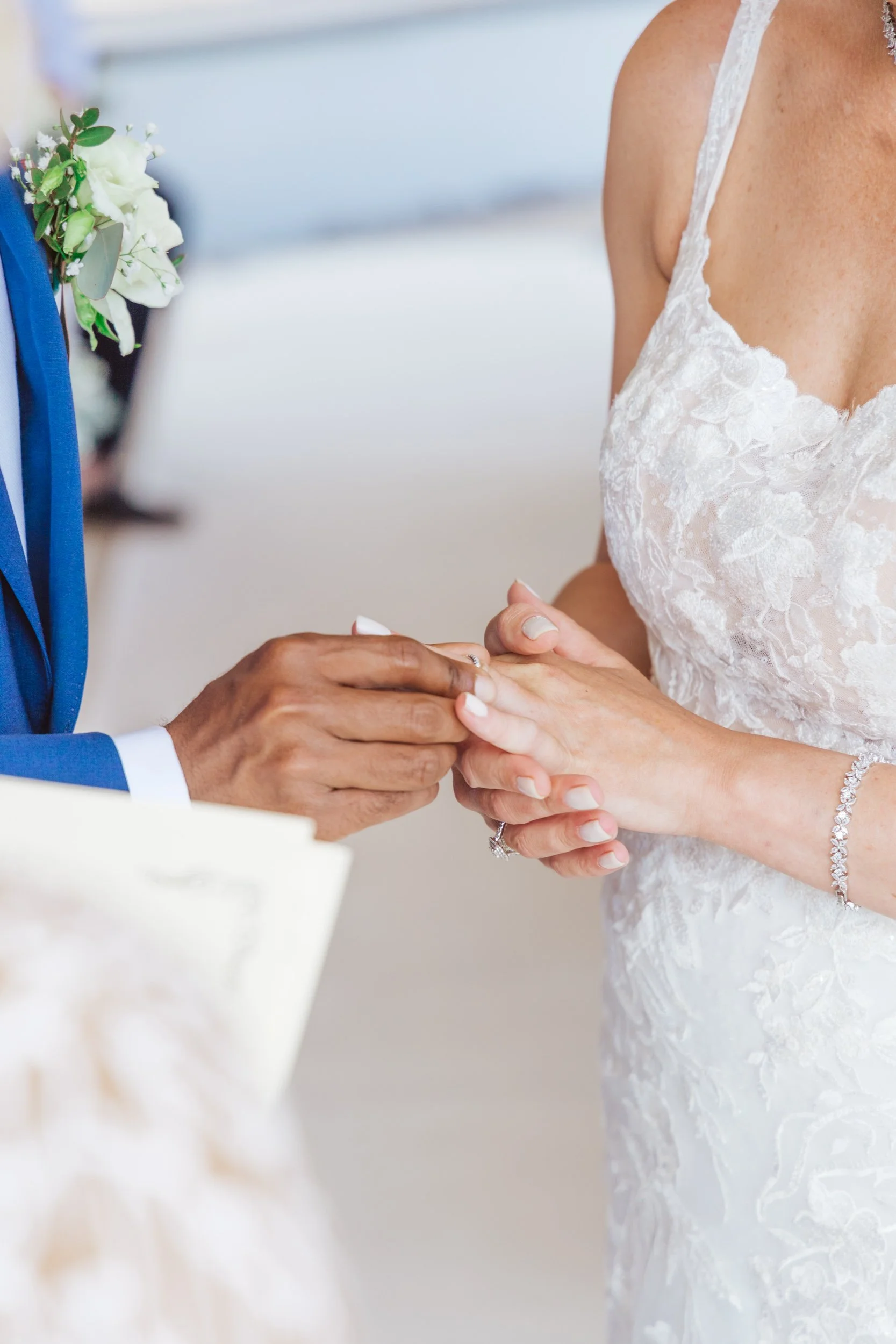 A couple is exchanging rings during a wedding ceremony, with the groom placing a ring on the bride's finger. The bride is wearing a white lace wedding dress and jewelry, and the groom is in a blue suit with a boutonniere.