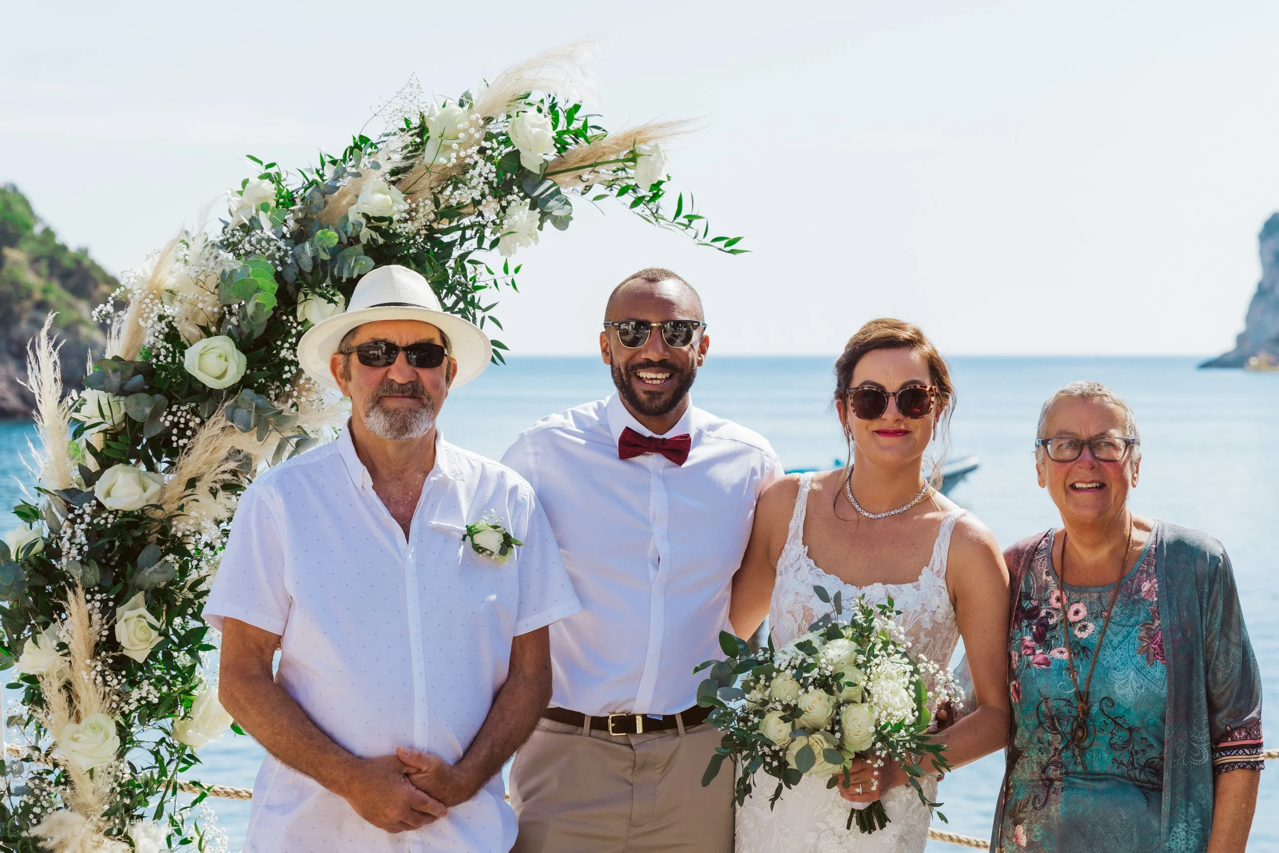 A wedding group photo with four people standing under a floral arch near the water. The bride in a white dress holds a bouquet, and a groom in a white shirt with a red bow tie stands beside her. Two older individuals, a man in a white shirt and woman