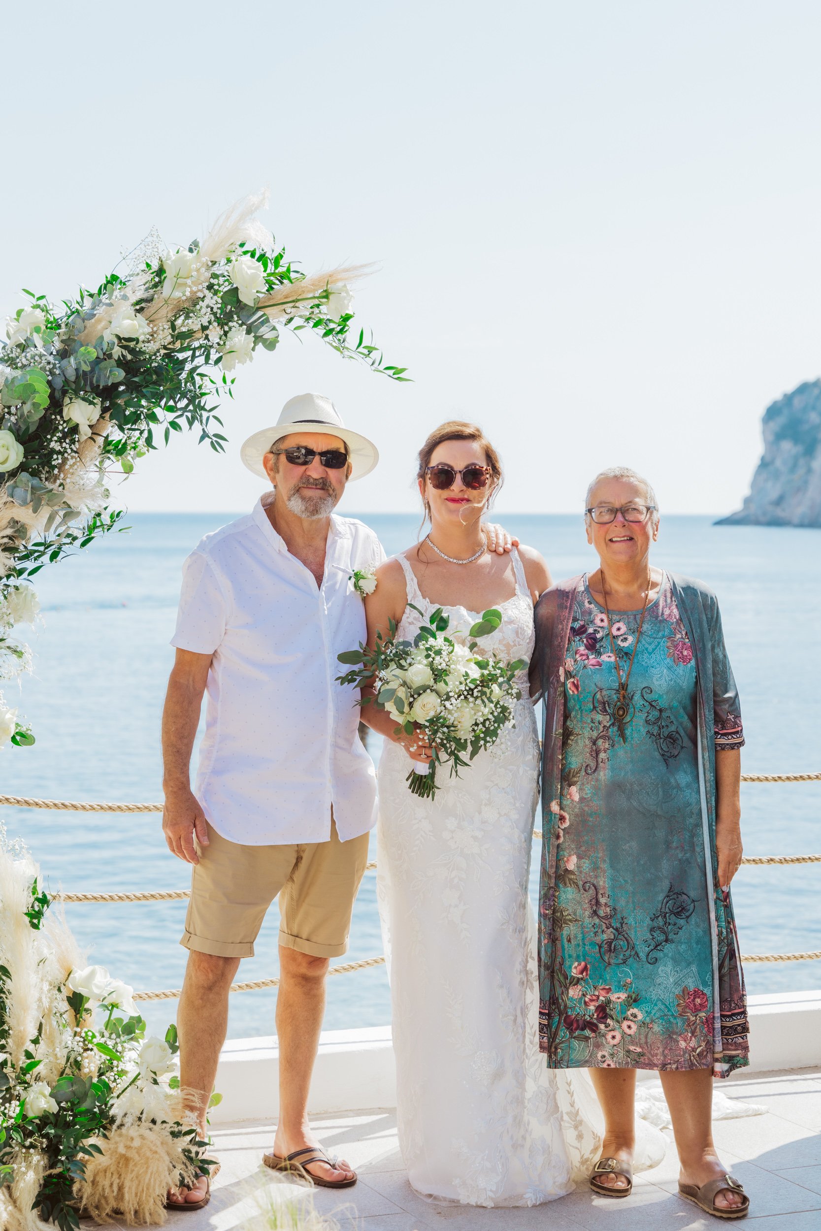 A bride in a white lace wedding dress holding a bouquet of white flowers, standing with an older man in a white shirt and shorts, and an older woman in a patterned dress, posing on a beachside wedding ceremony with an ocean and rocky cliff in the bac