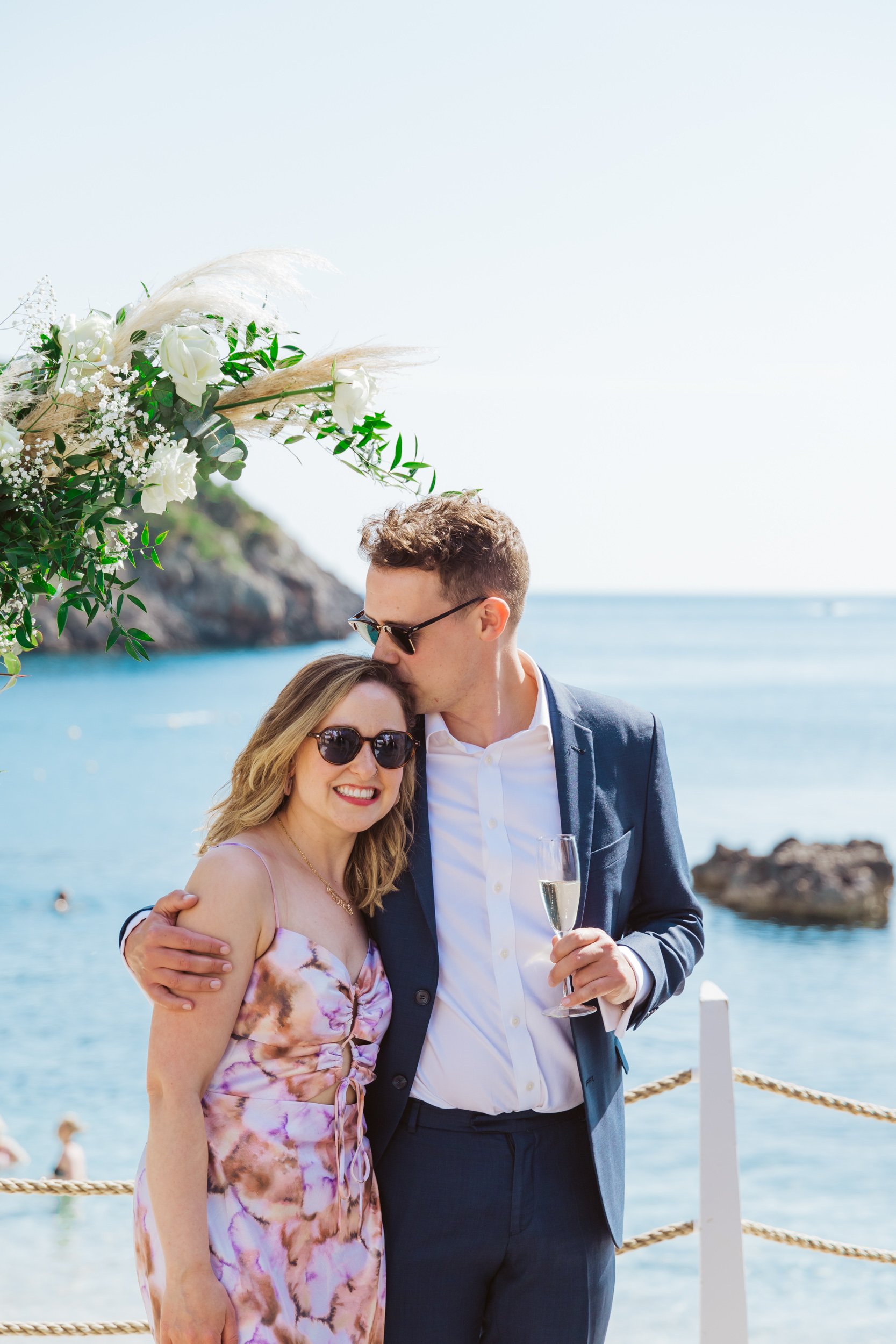 A couple dressed in semi-formal attire, with the man in a navy suit and the woman in a colorful floral dress, standing together by the beach with the man kissing the woman's forehead, during daytime with a blue sky and water in the background.