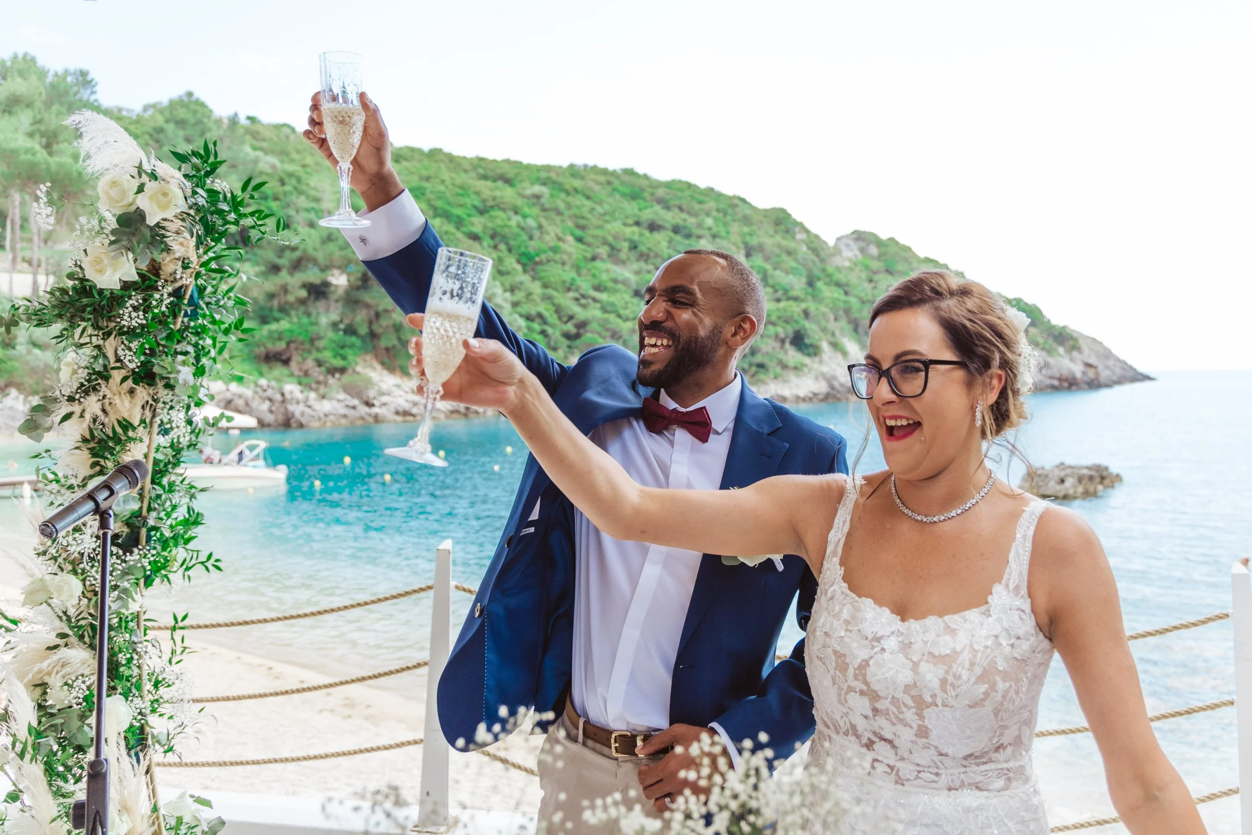 A newlywed couple celebrating with champagne beside a decorated arch at a seaside wedding altar, with a scenic ocean view and green hills in the background.