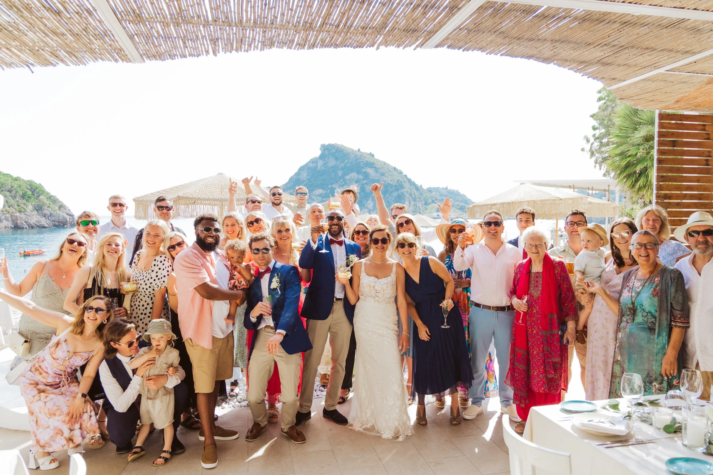A large group of people at a wedding celebration on a beachside venue, with mountains in the background, under shaded cover, featuring men and women of various ages dressed in formal and casual attire, some holding glasses and children present.