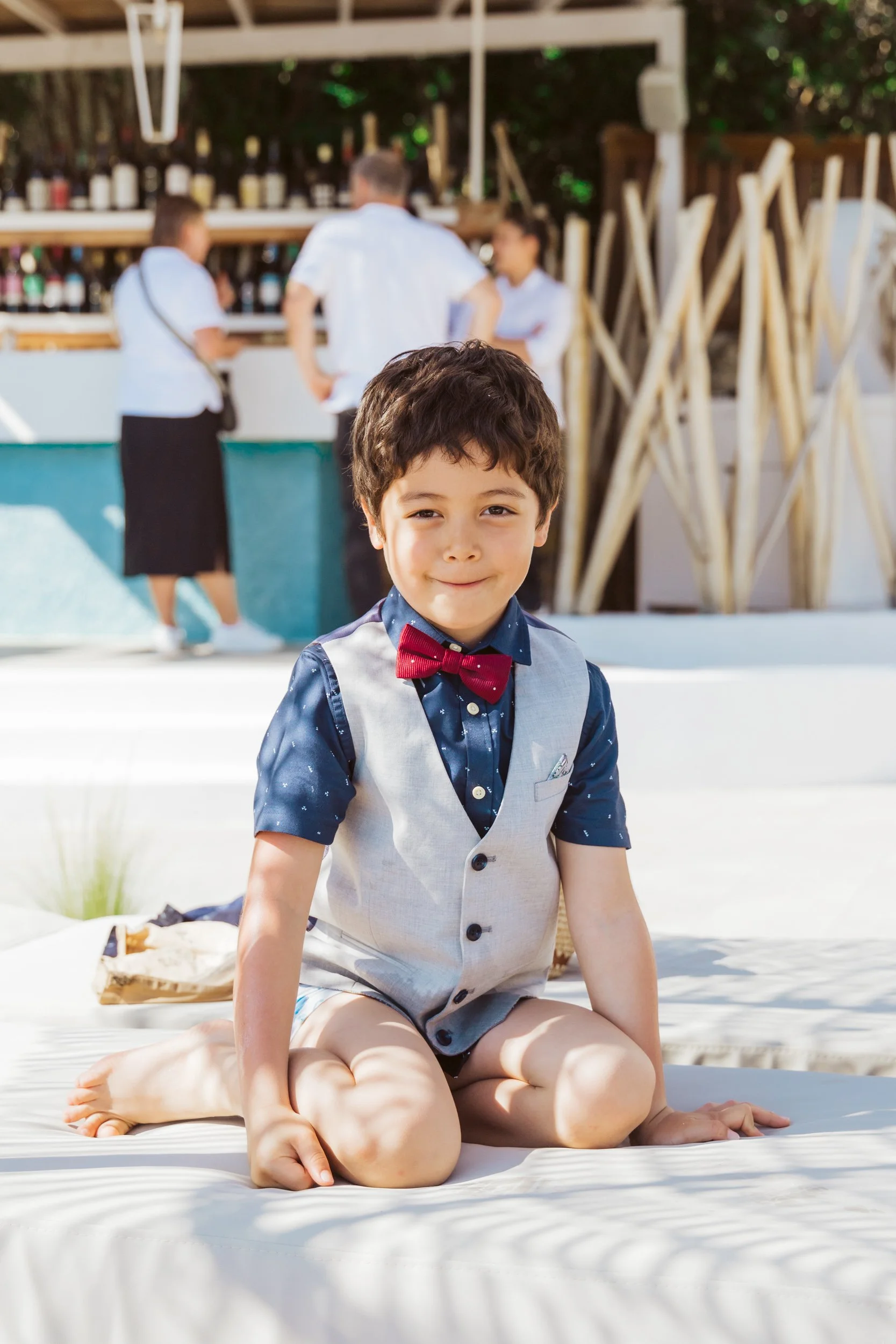 Young boy in formal attire with a red bow tie sitting on his knees on a white outdoor lounge chair, smiling at the camera, with a bar and three people in the background.