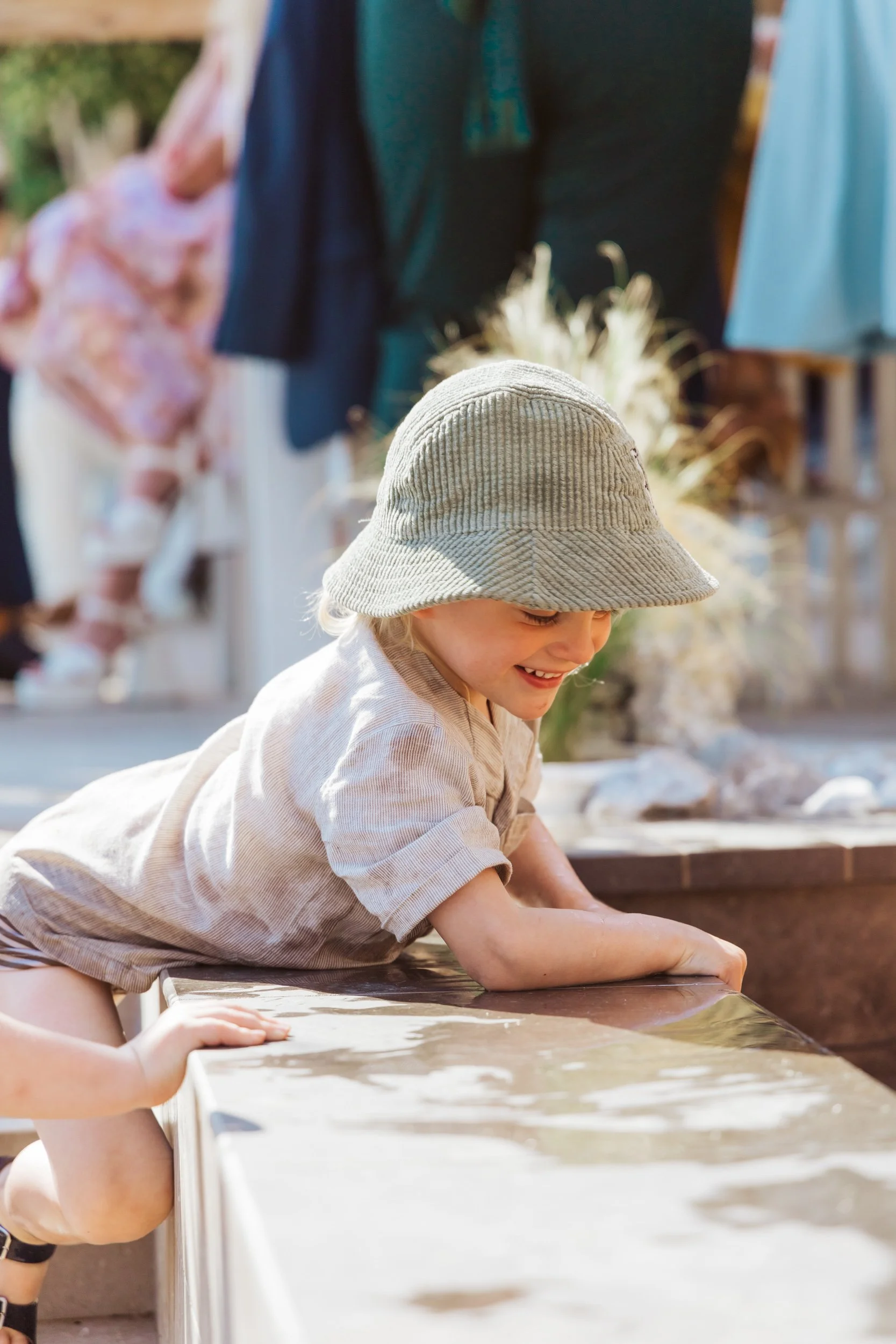 A young boy in a beige hat and shirt leaning over a water table, smiling and playing outdoors.