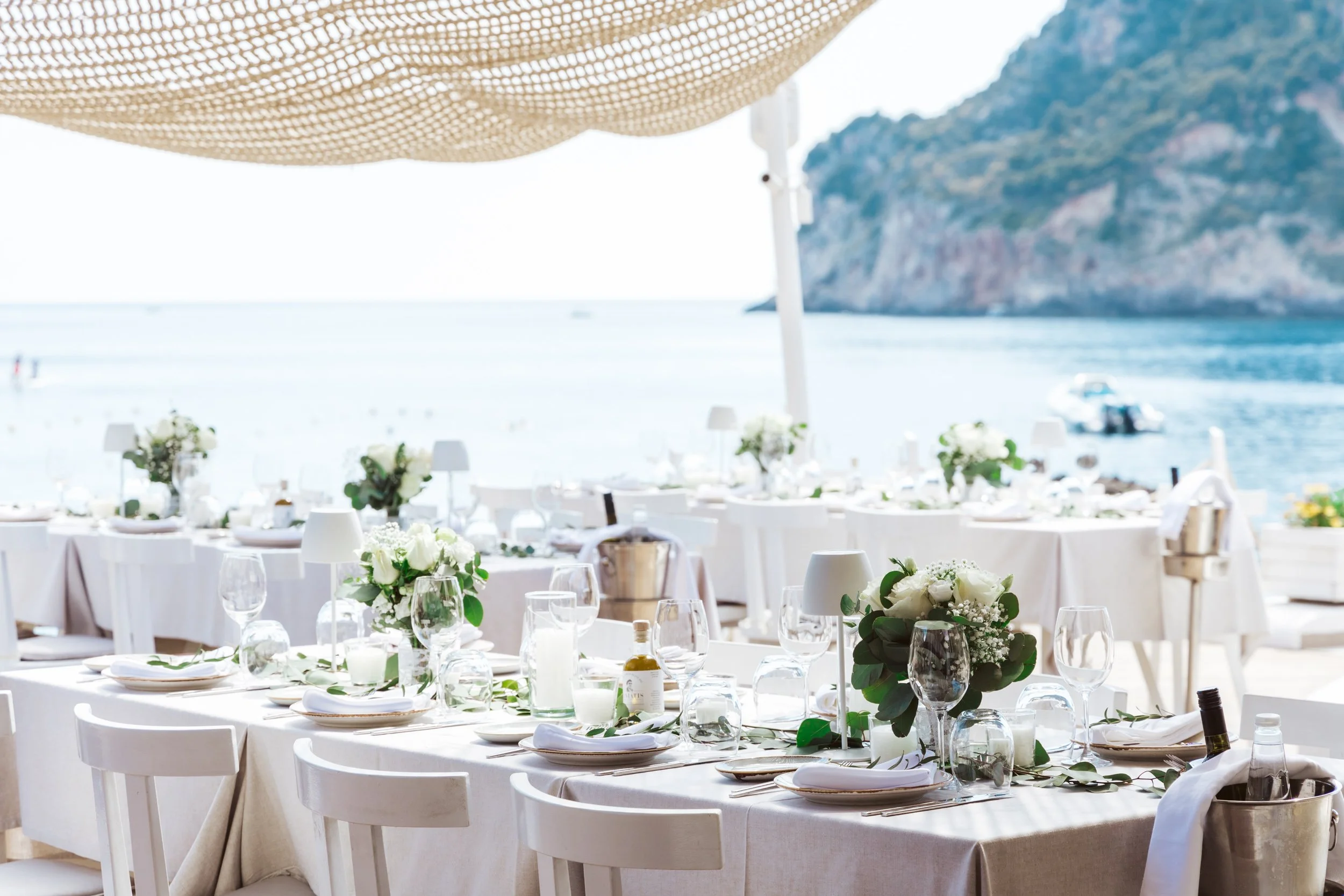 Elegant beachside banquet setup with white tablecloths, floral centerpieces, and tableware, overlooking the ocean and rocky cliffs.
