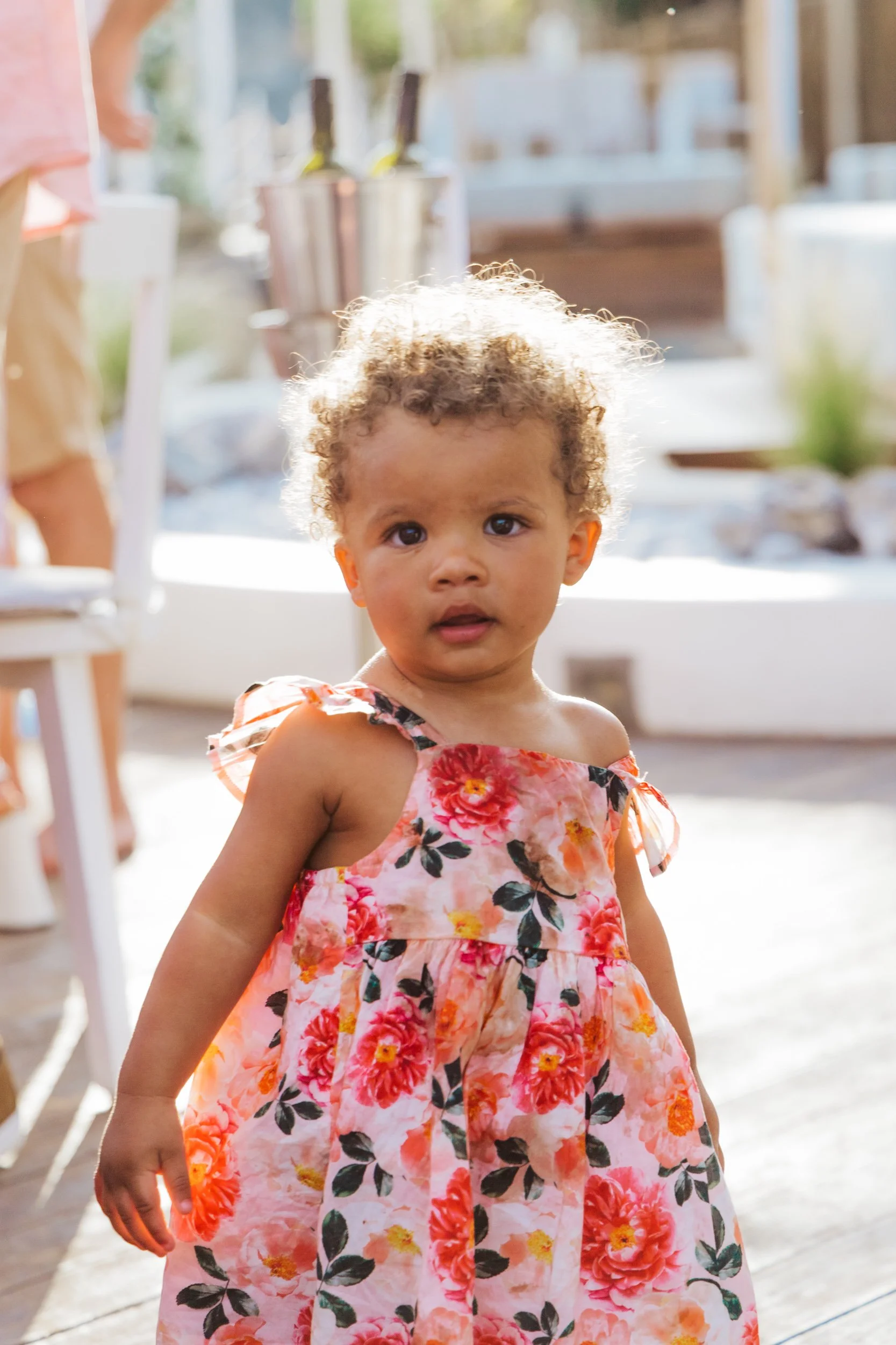 A young girl with curly hair wearing a floral dress, standing indoors with sunlight in the background.