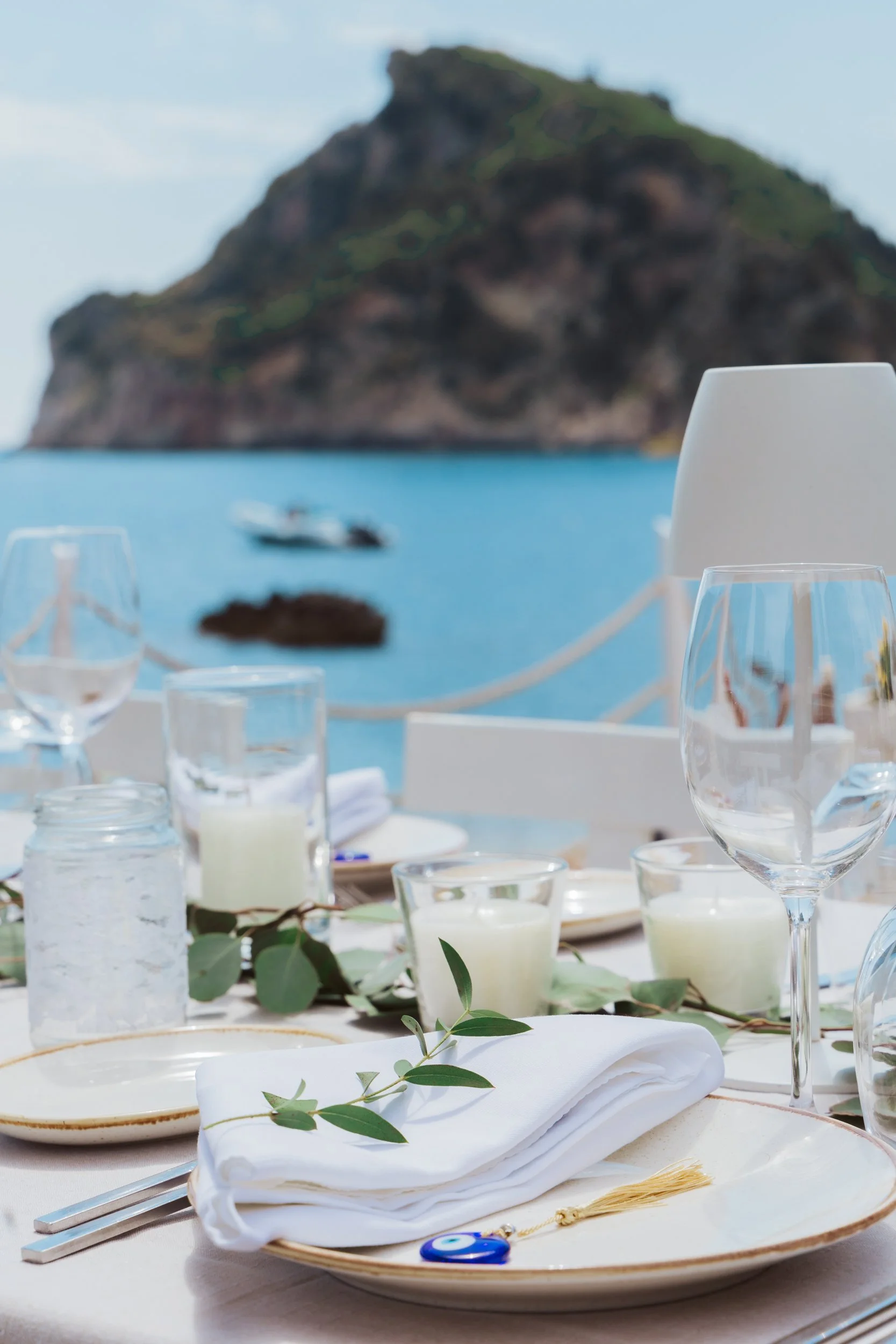 Elegant outdoor table set with white plates, green leaves, glasses, candles, and a dreamy ocean view with a distant island mountain.