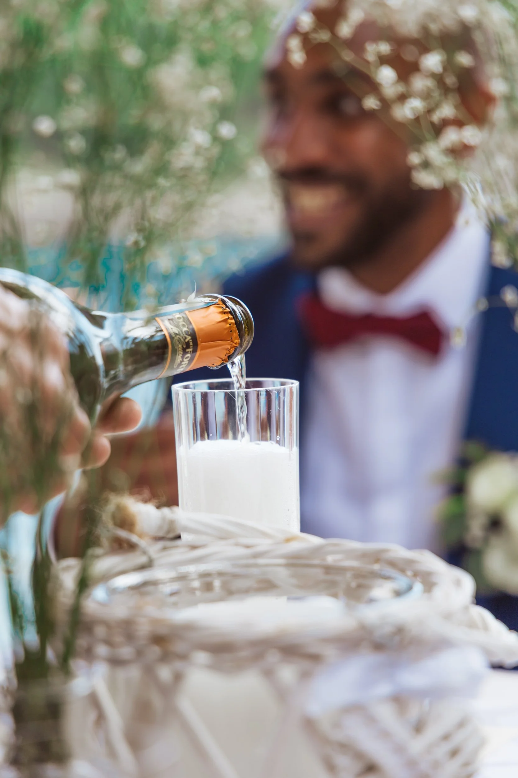 A man in a blue suit and red bow tie smiling as champagne is poured into a glass on a wedding cake table.