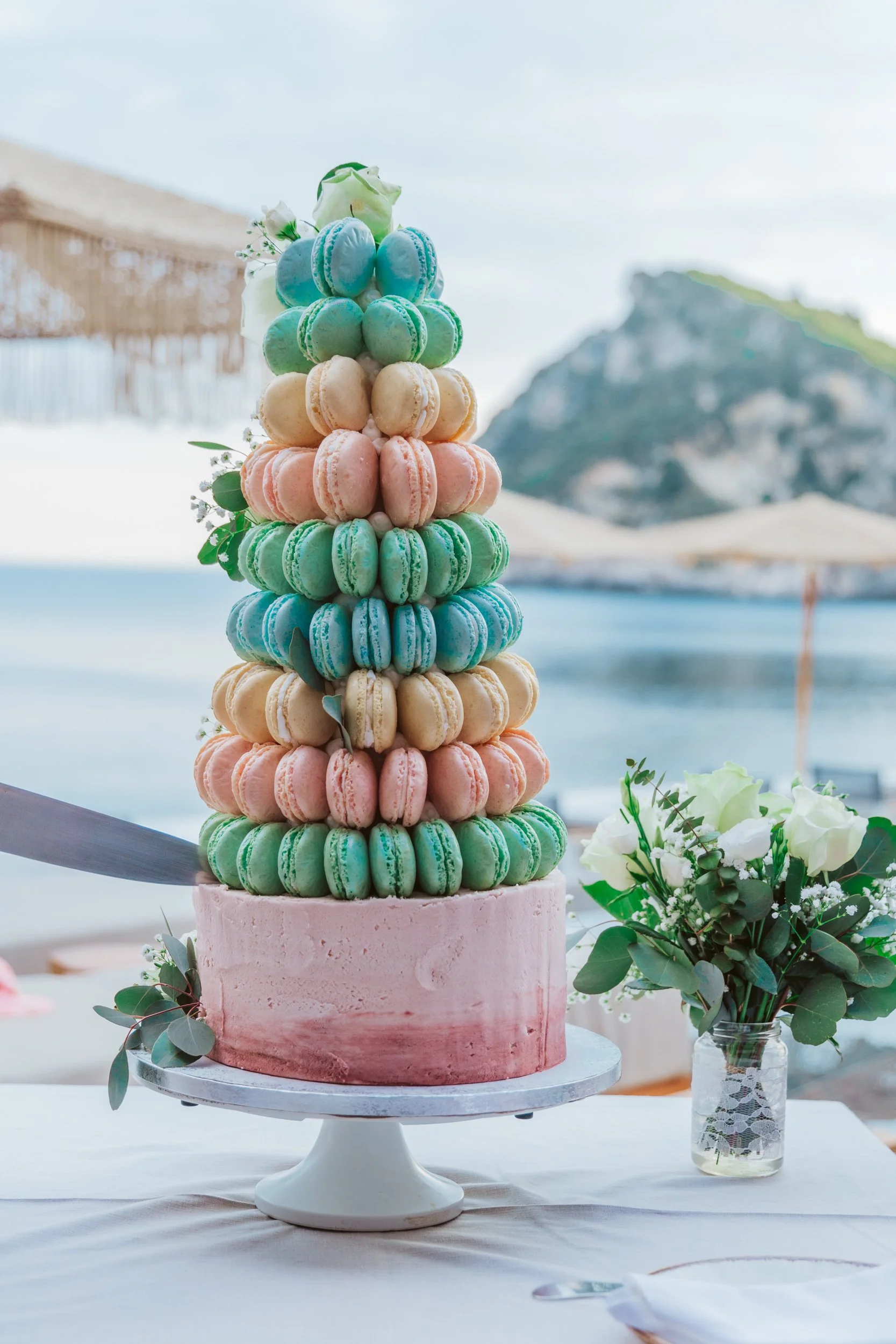 A colorful macaroon tower with pastel pink, green, blue, and beige macarons, decorated with white roses and greenery, on a cake stand at a beachside setting.