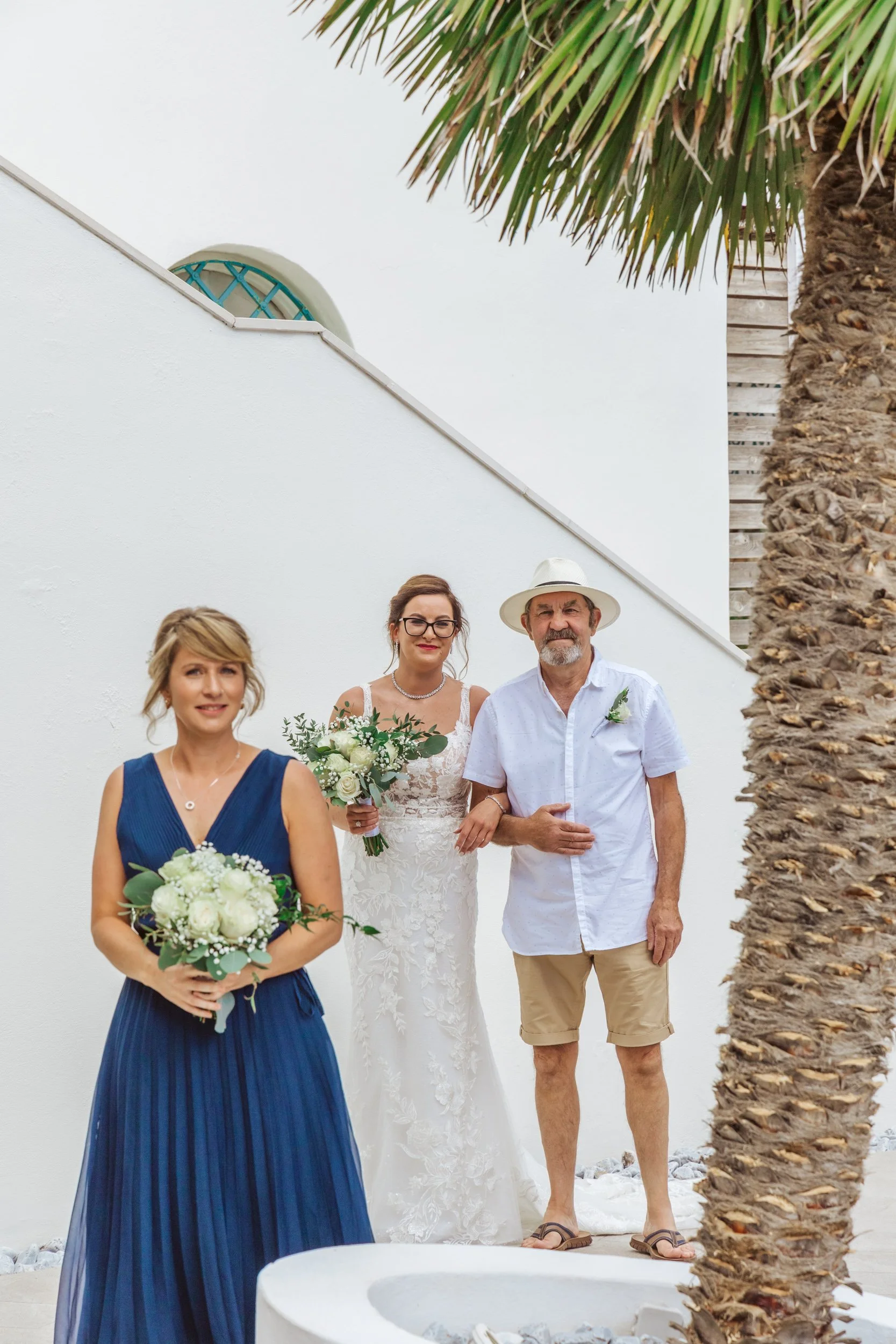 A woman in a blue dress holding a bouquet, a bride in a white wedding dress holding a bouquet, and an older man in a white shirt, beige shorts, and a white hat standing together outdoors with a white wall and a palm tree.