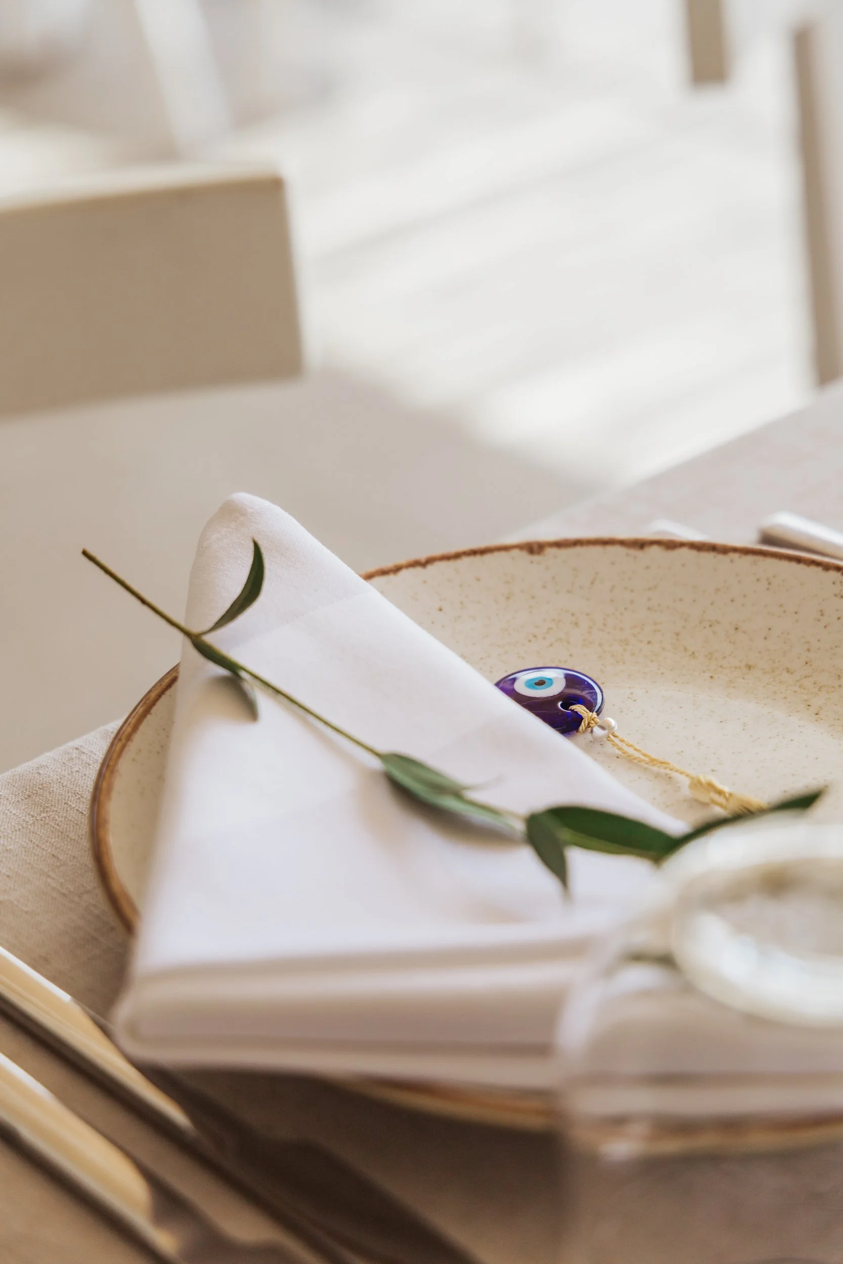 A white cloth napkin folded on a speckled beige plate with a small green leafy branch on top. There is a blue evil eye charm nearby on the plate. The place setting is on a light-colored table.