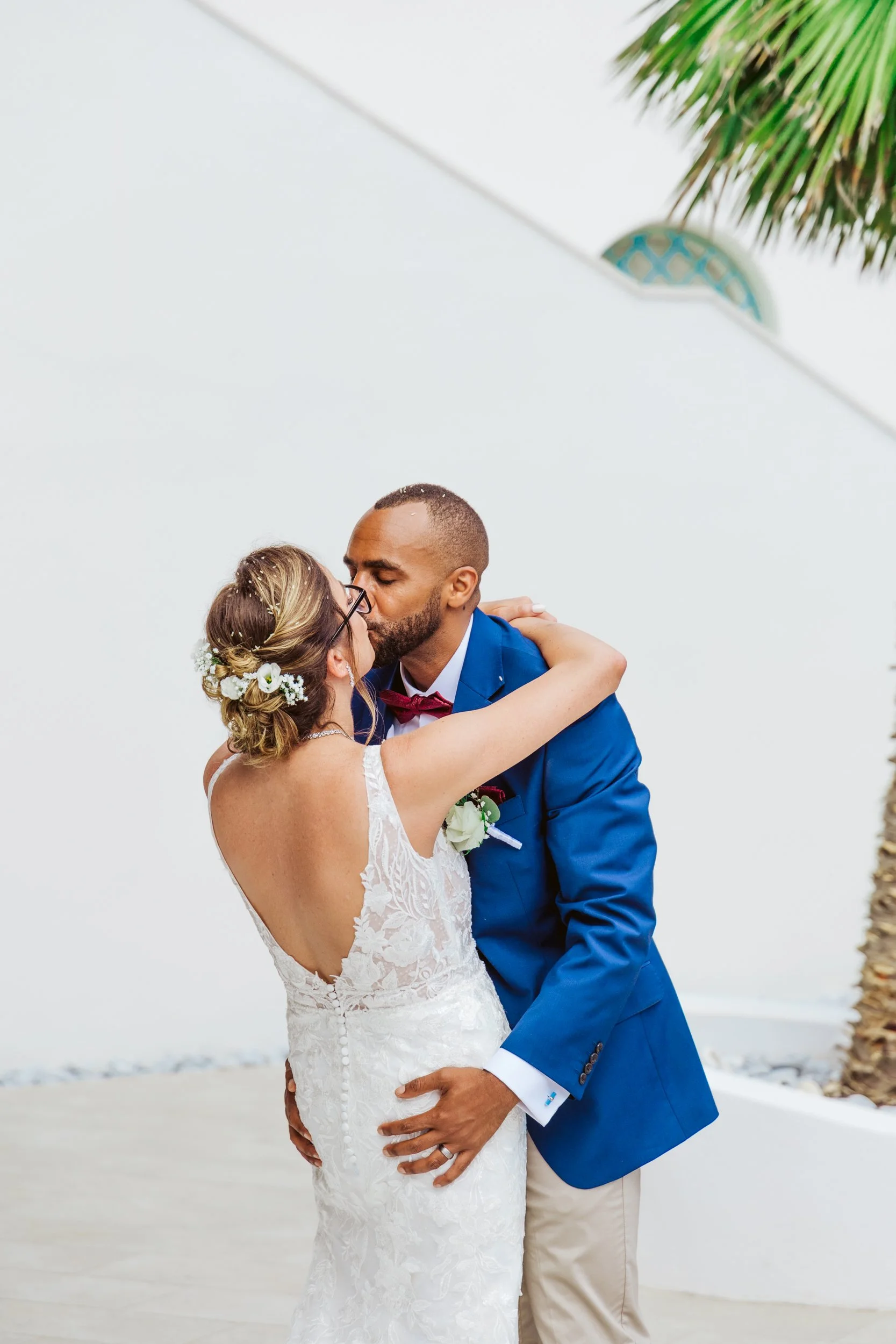 A newlywed couple sharing a kiss outside, the bride with floral hair and glasses, the groom in a blue suit and red bowtie, against a white wall and some tropical plants.