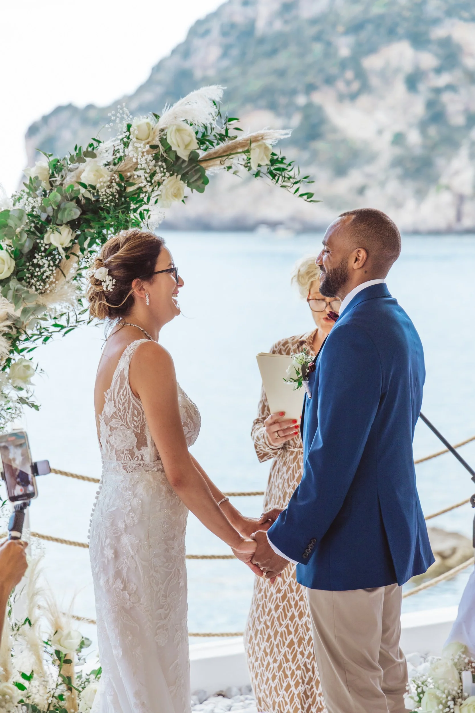 A wedding ceremony taking place outdoors by the water, with a bride and groom holding hands and facing each other, under a floral arch. An officiant is standing behind them, and a person is recording the ceremony with a phone.