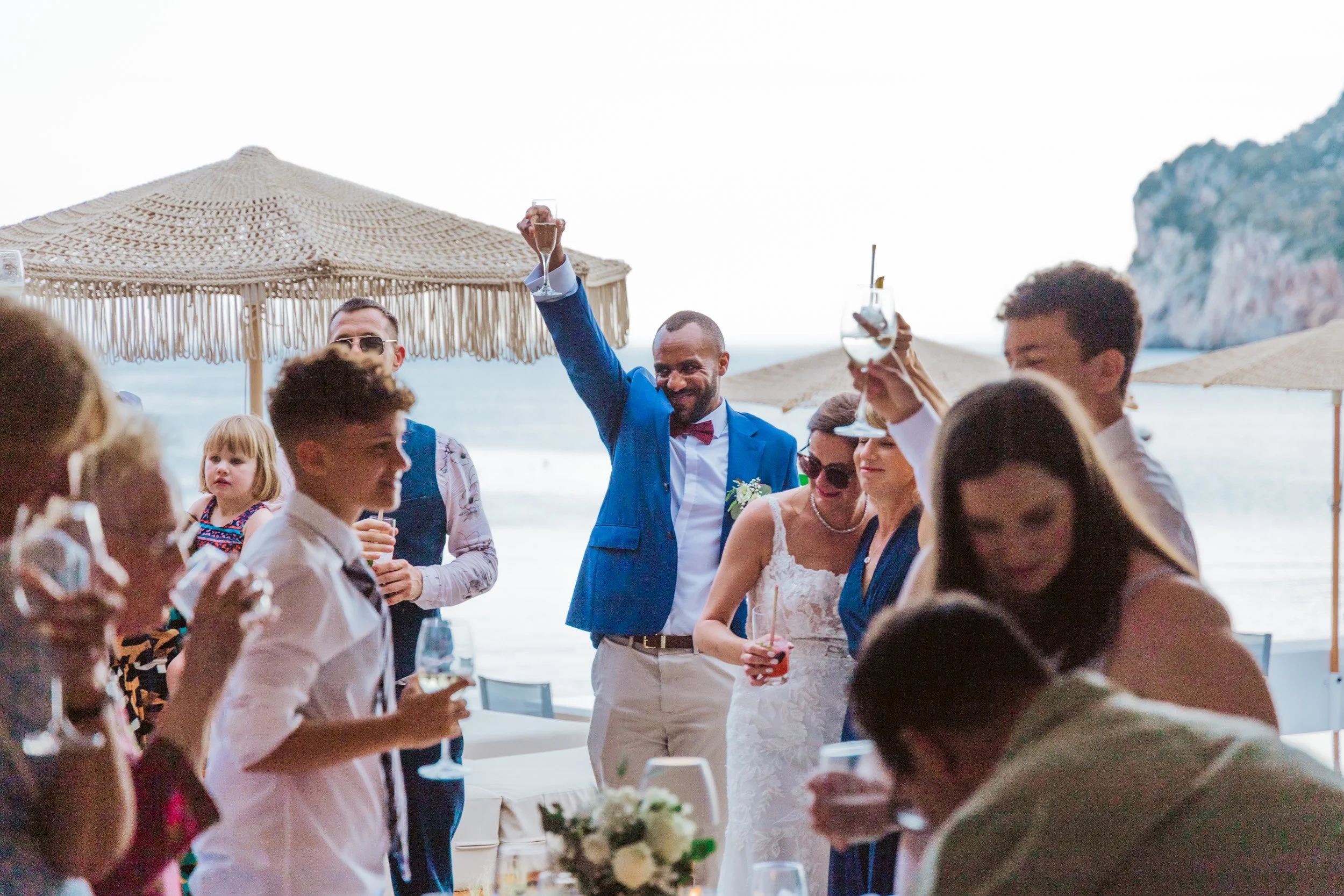 Group of people celebrating at a beach wedding reception, with some raising glasses in a toast, umbrellas and ocean in the background.