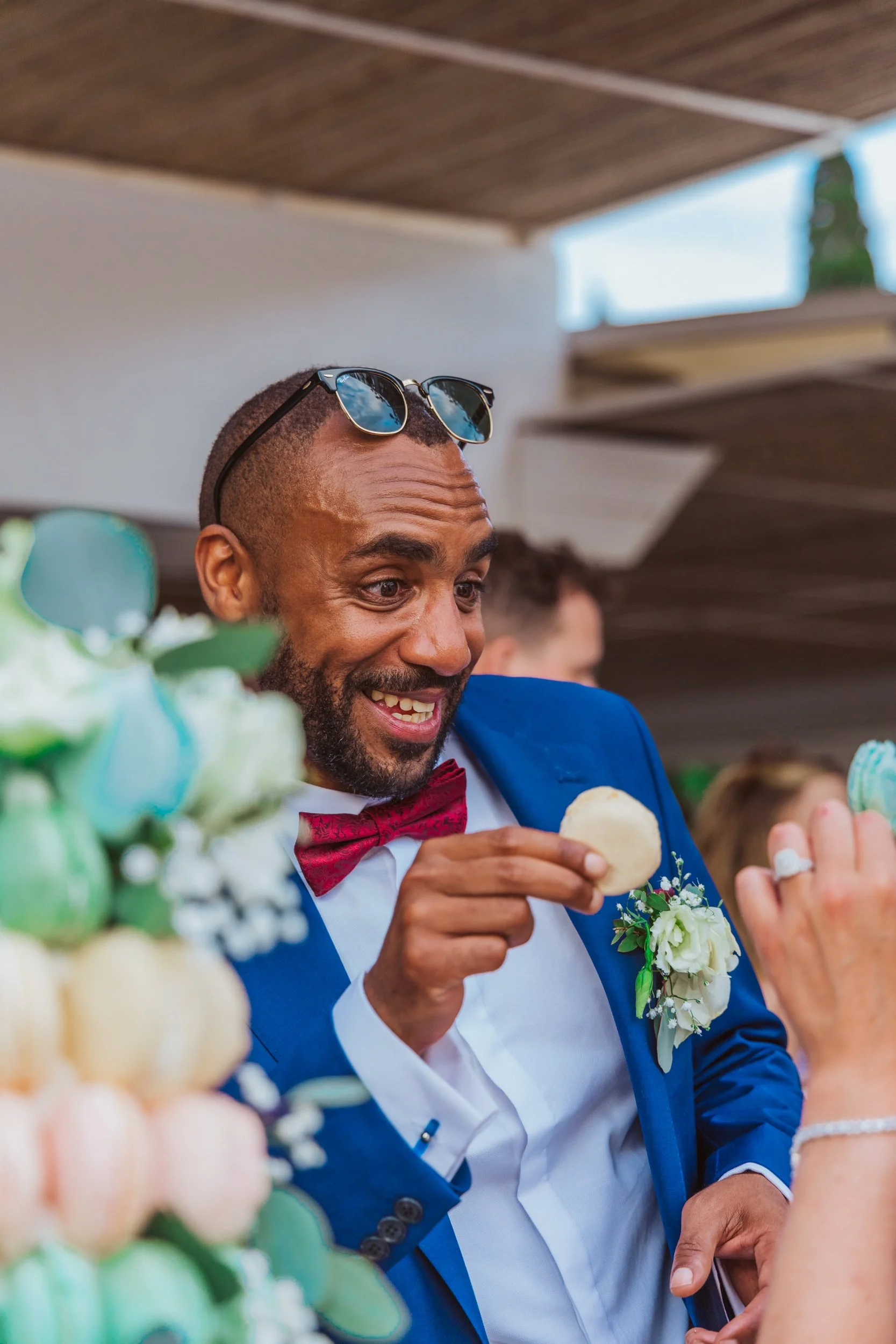 A man with sunglasses on his head, wearing a blue suit and red bow tie, smiling and enjoying a macaroon while at a celebration, with blurred people and a wooden ceiling in the background.