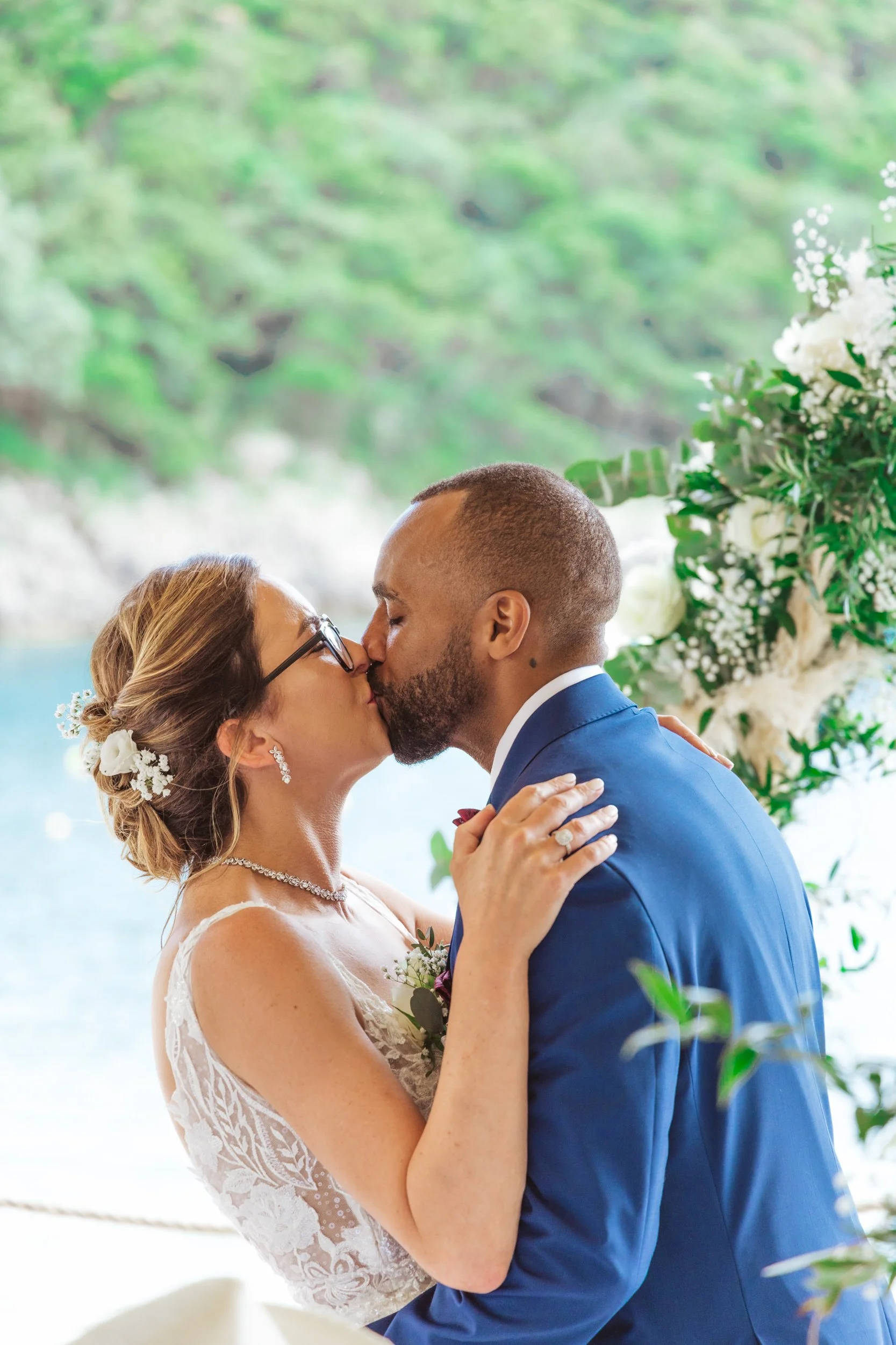 A bride and groom kiss during their wedding ceremony with greenery and flowers in the background.