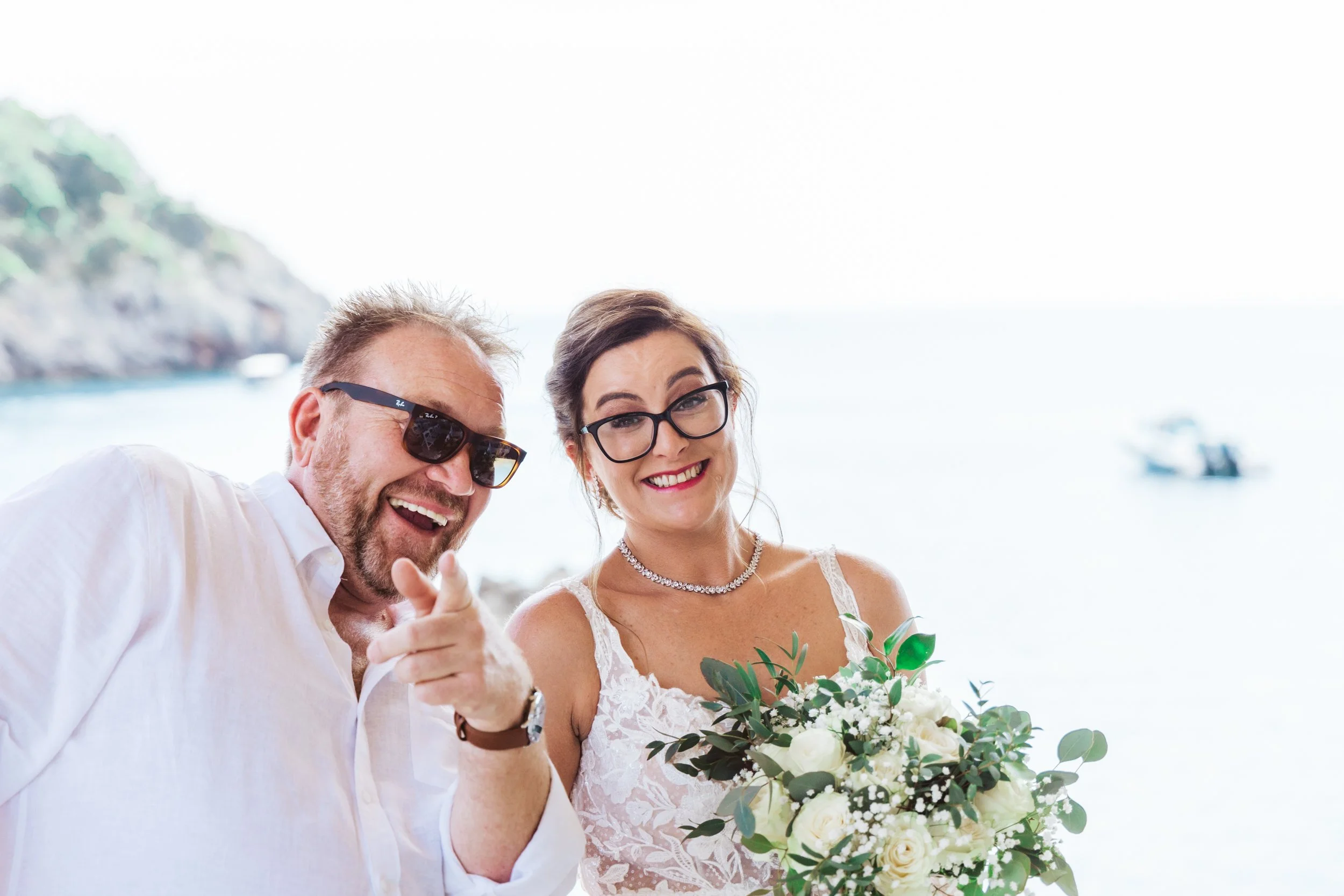 A happy couple at a wedding, with the woman holding a bouquet of white roses, standing outdoors near water with boats in the background.