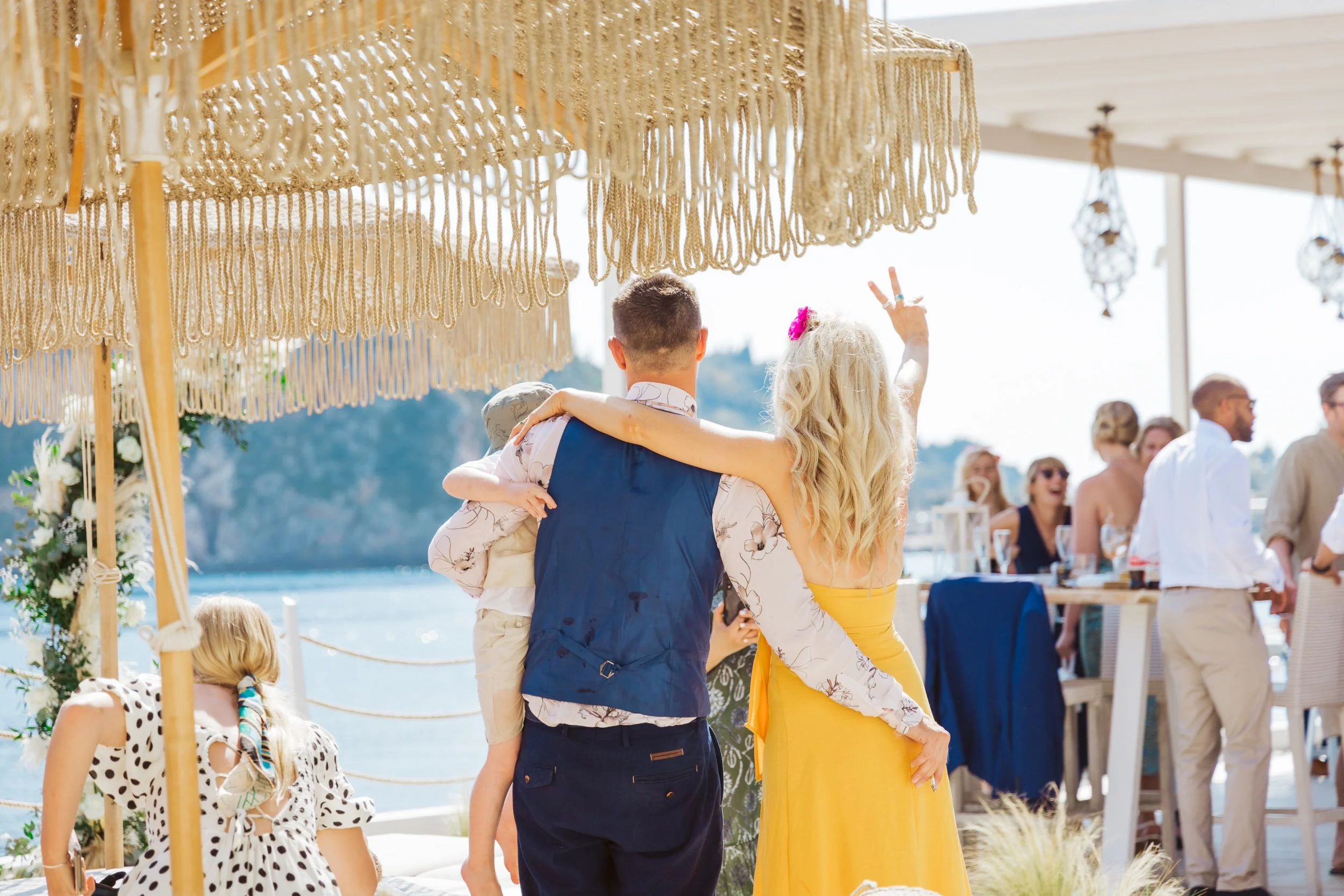 A group of people celebrating at an outdoor event near the water on a sunny day. A woman with long blonde hair in a yellow dress is being held up by a man in a vest, with a child on his back. Other guests are standing and sitting nearby under decorat