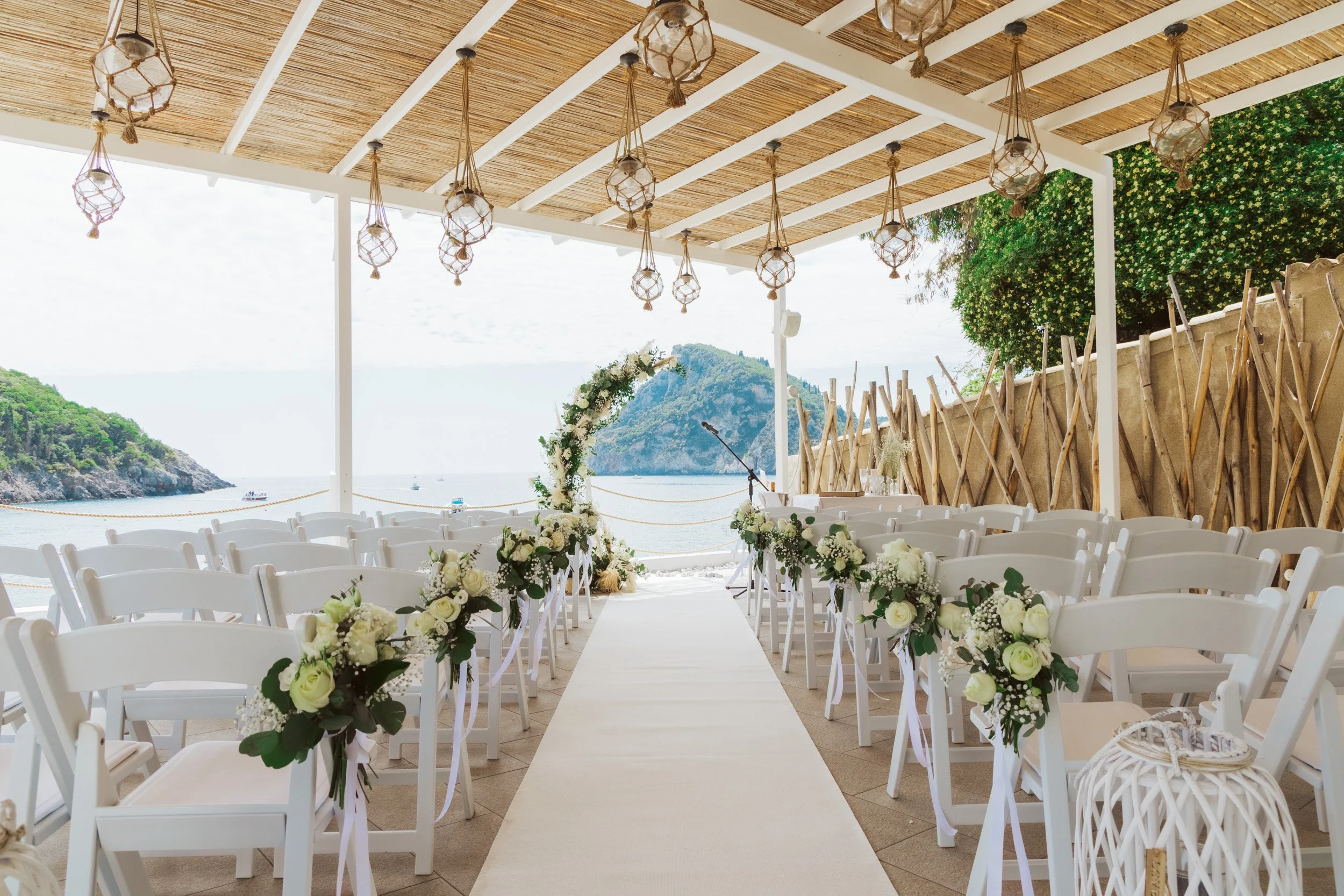 An outdoor wedding ceremony setup with white chairs decorated with white flowers along an aisle, facing an ocean view with rocks and green hills in the background. There is an arch decorated with flowers, and the space is covered with a wooden roof a