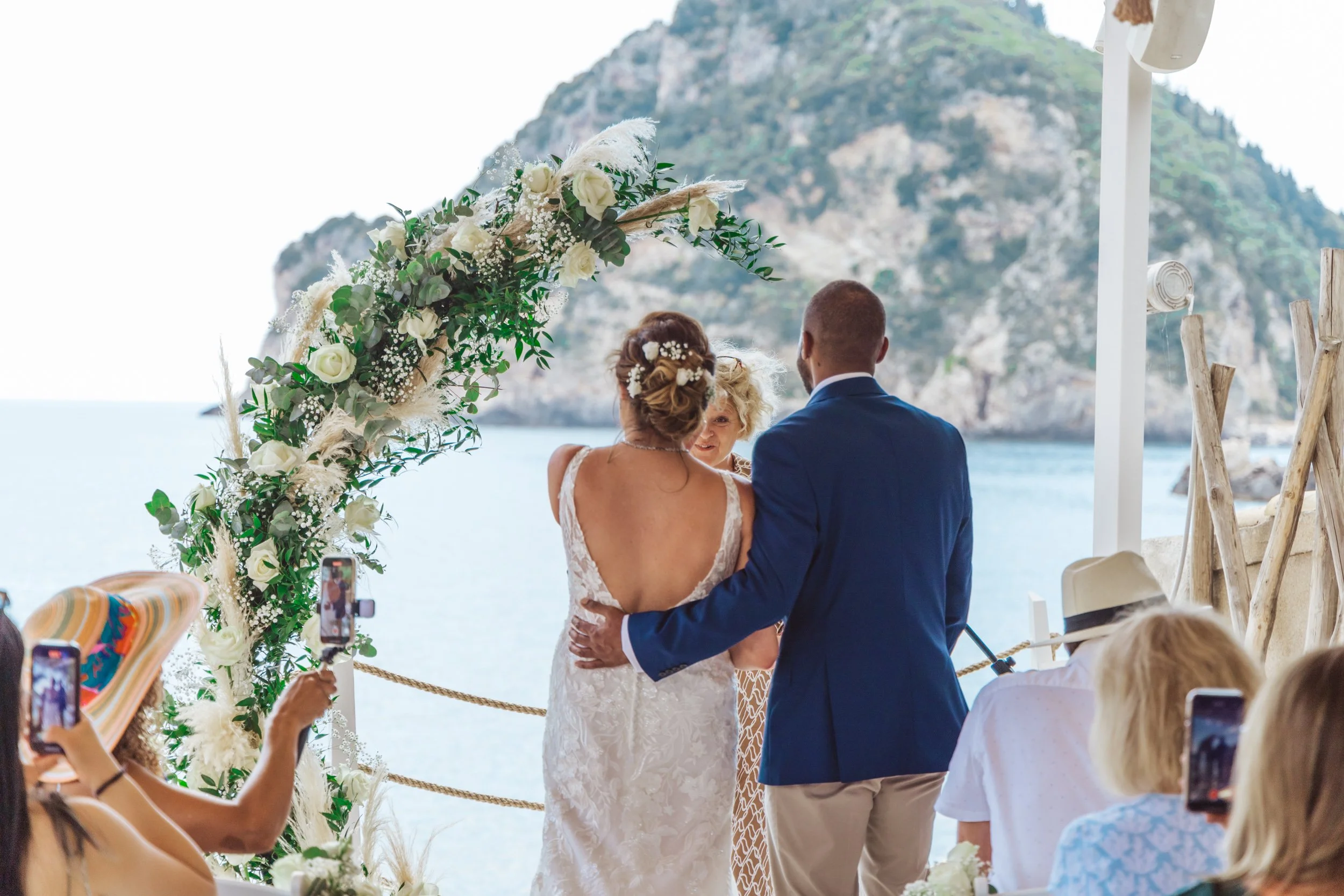 A wedding ceremony taking place outdoors by the water with a scenic mountain backdrop, featuring a bride in a lace wedding dress and a groom in a blue suit, standing under a floral archway, surrounded by seated guests capturing the moment on their ph