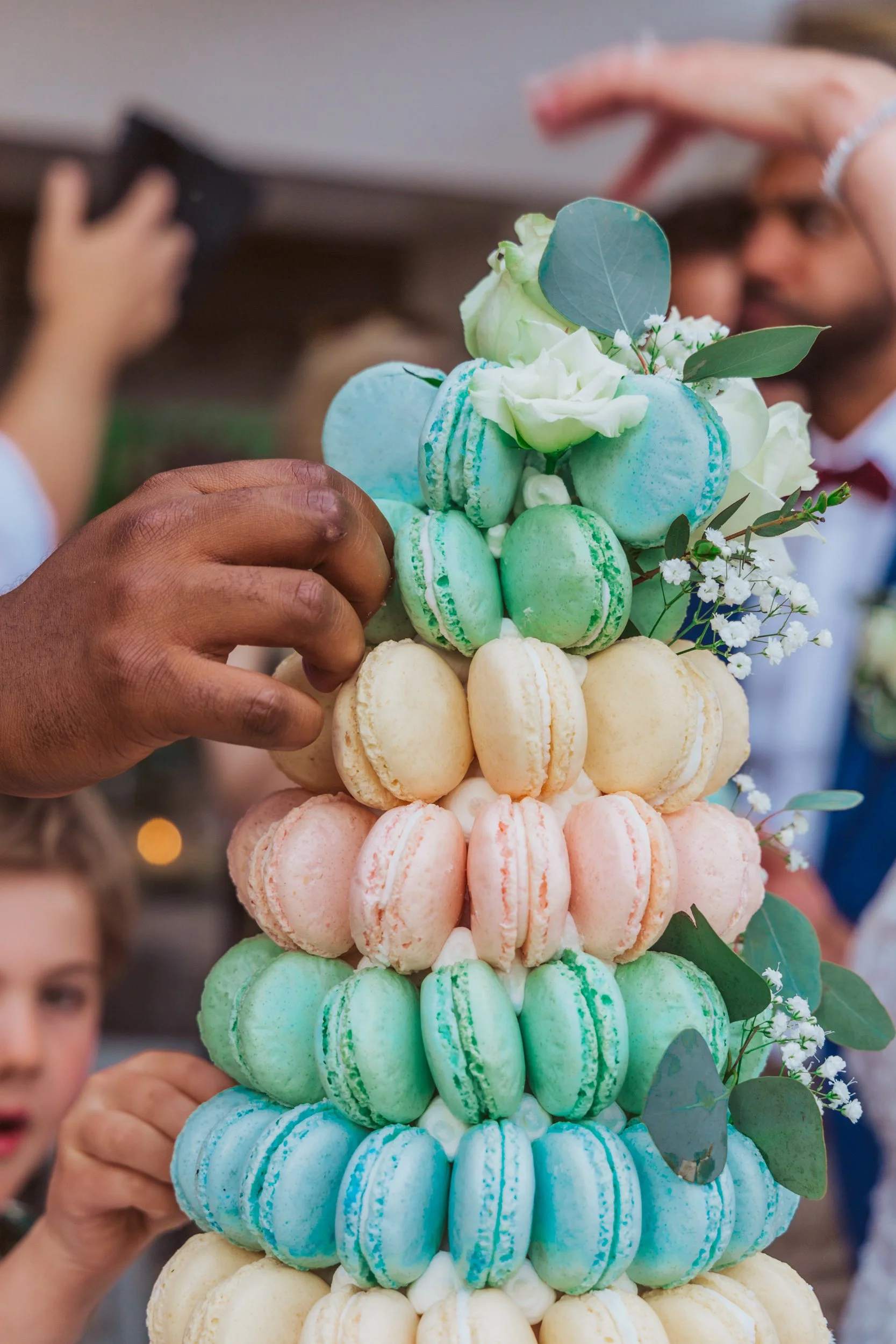 A colorful tower of macarons with flowers and eucalyptus leaves on top, during a celebration.