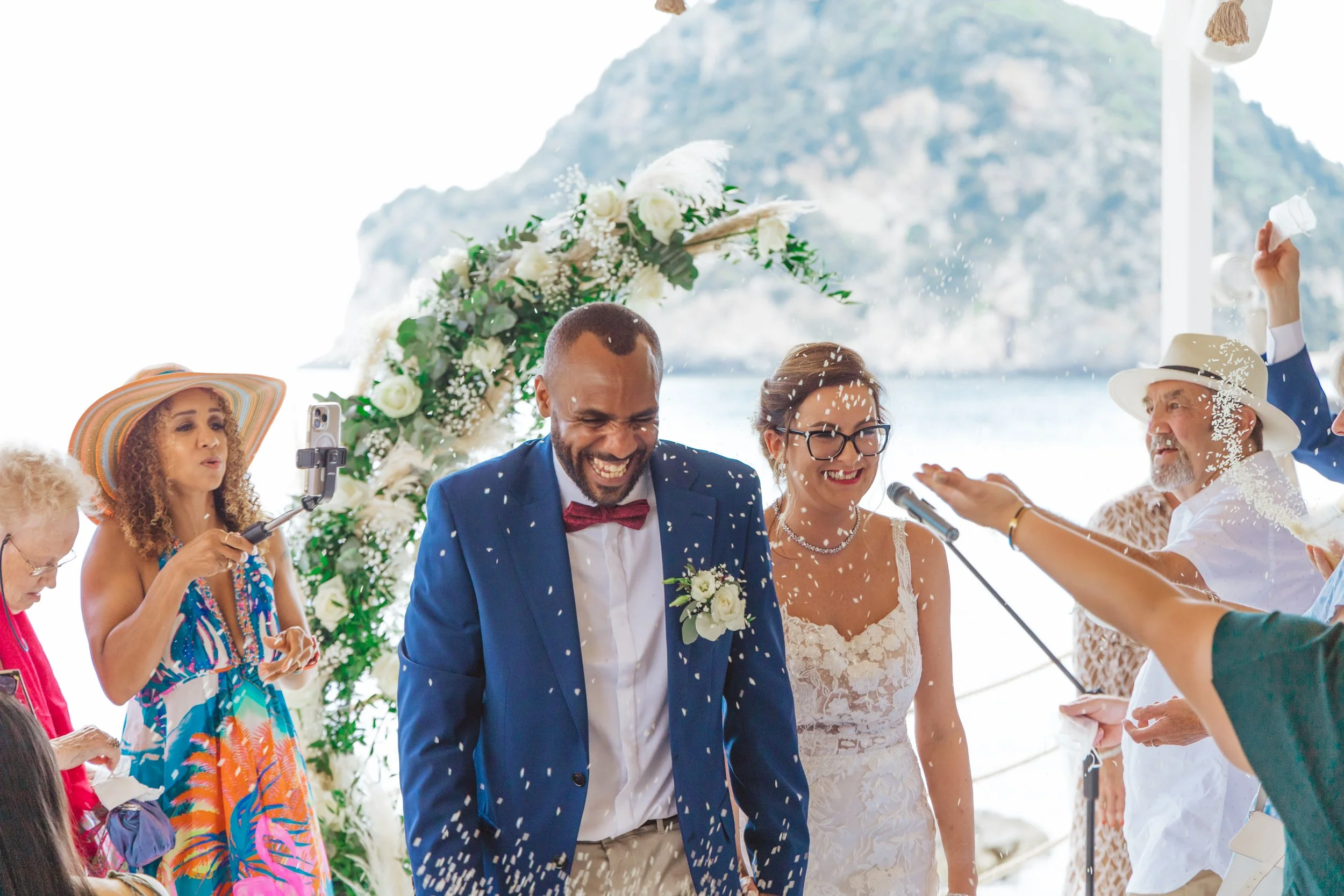 A newlywed couple smiling during their outdoor wedding ceremony by the water, surrounded by family and friends, with white floral arrangements and an ocean view in the background.