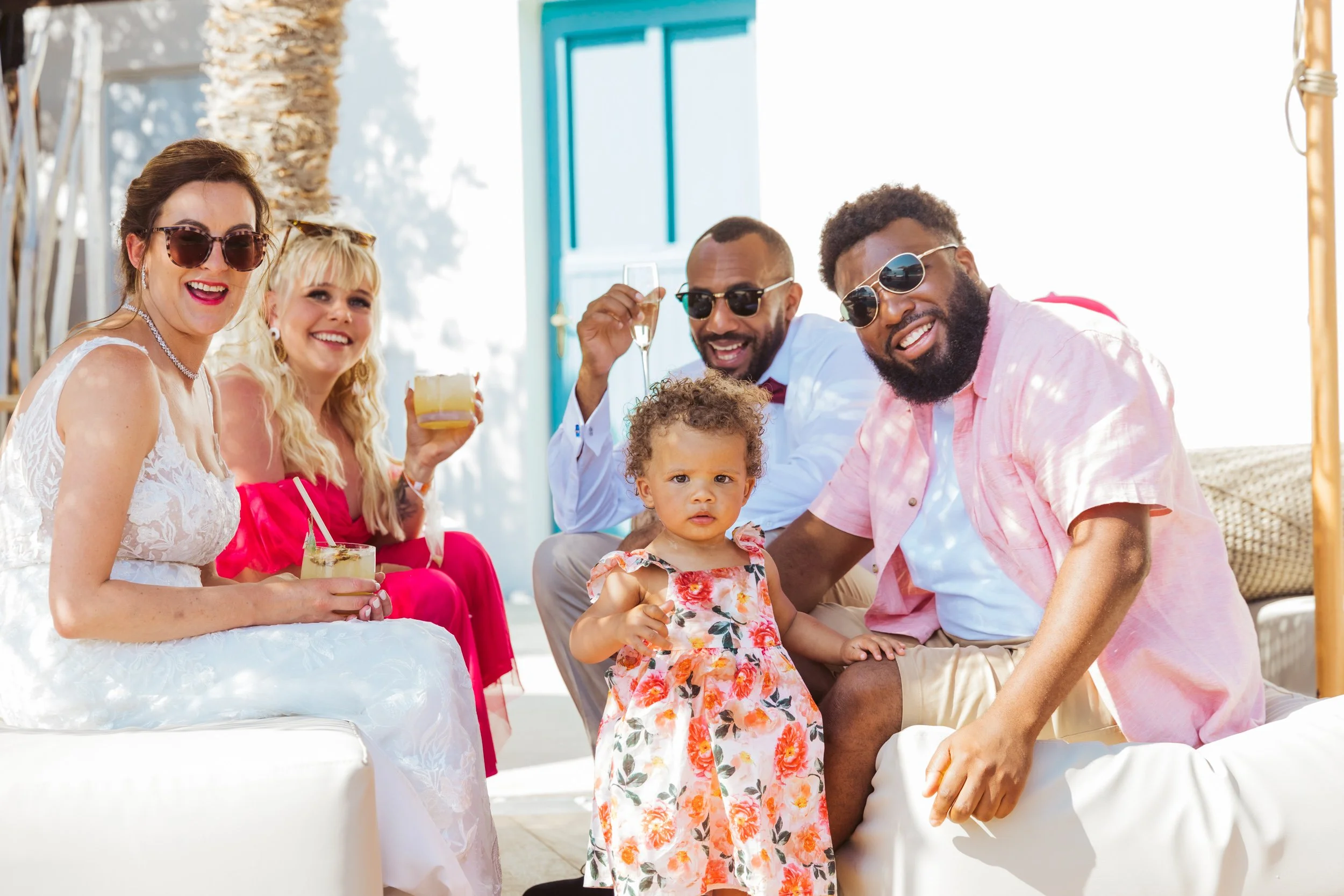 Group of six people sitting together outdoors at a party, dressed in summery clothes, some holding drinks, with a young girl in the foreground, all smiling or looking at the camera, with a palm tree and bright sunlight in the background.