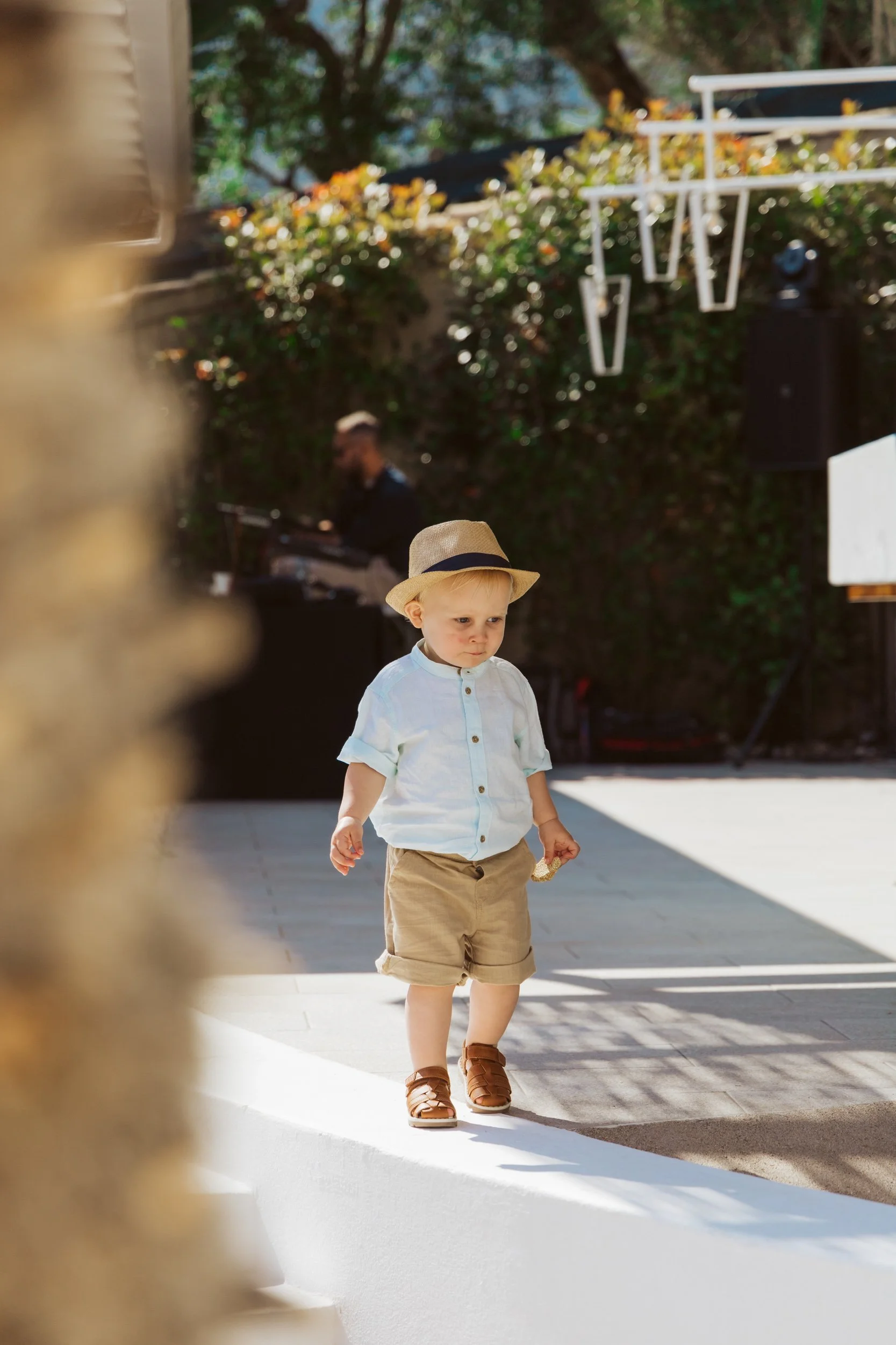 A young boy wearing a beige hat, white shirt, khaki shorts, and brown sandals walking outdoors on a bright sunny day. A musician playing keyboard is seen in the background.