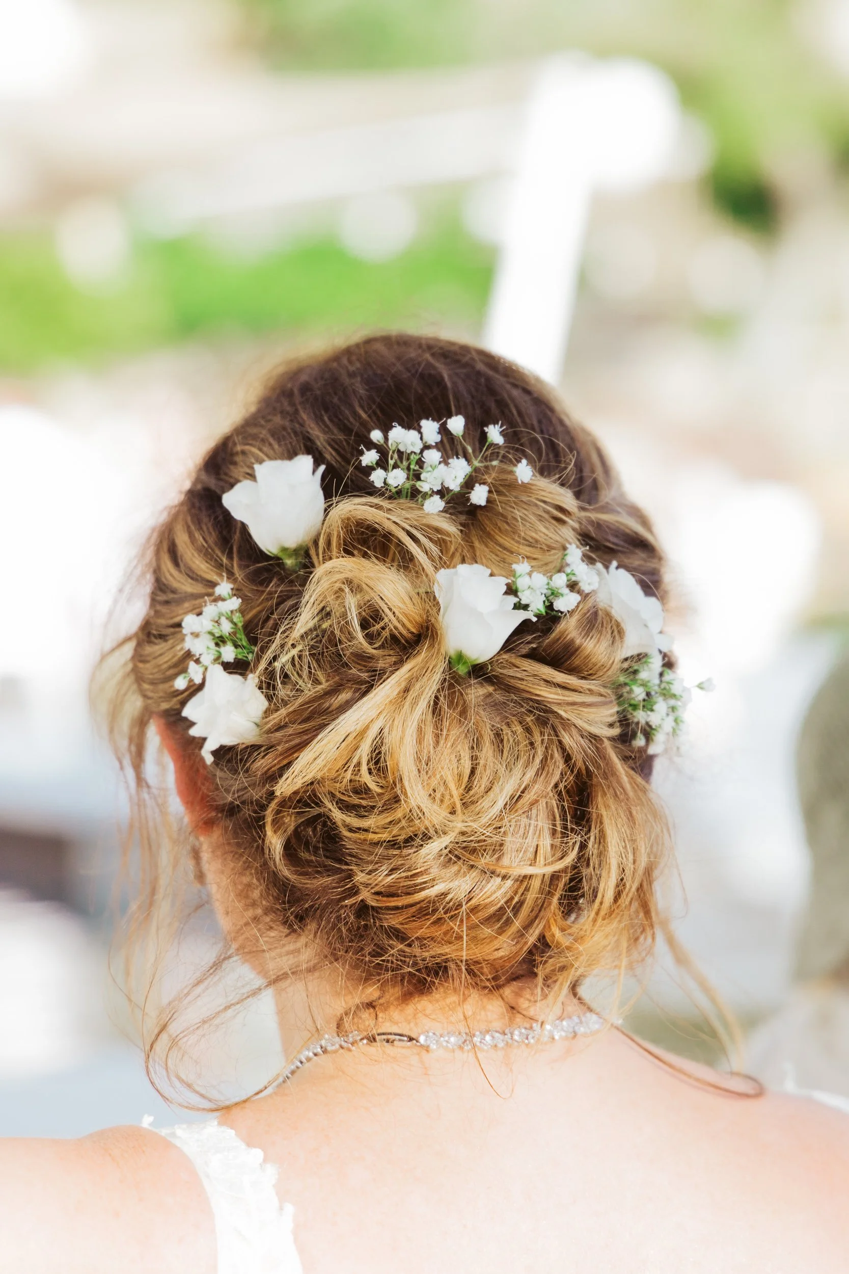 Back view of a woman with curly, blonde hair styled in an elegant updo adorned with white flowers and baby's breath. She is wearing a pearl necklace and a white lace dress, with a blurred outdoor background.