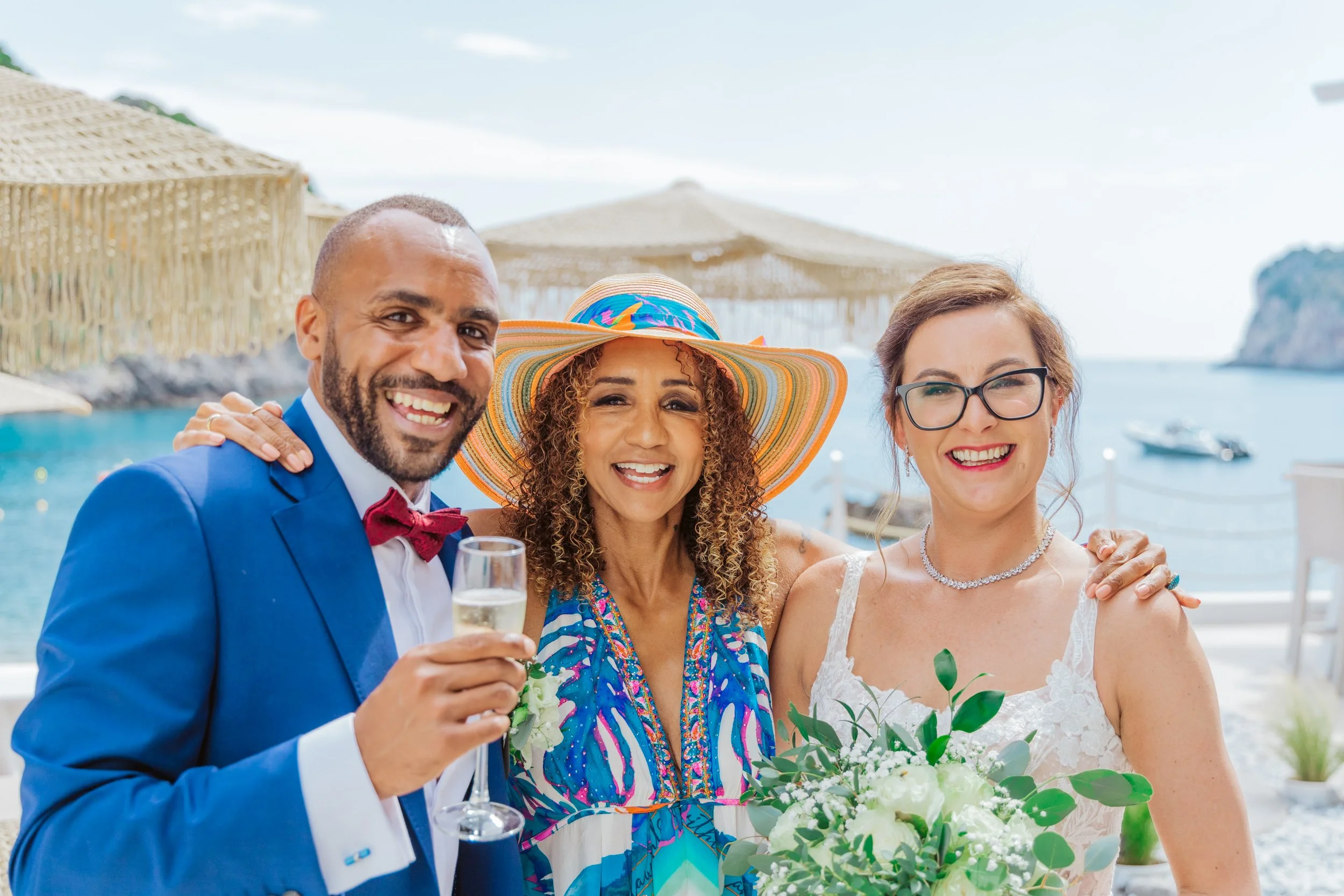 Three people at a beach wedding celebration, smiling and dressed in formal and beach attire, with a man in a blue suit holding a champagne glass, a woman in a white wedding dress holding a bouquet, and another woman in a colorful dress and large sunh