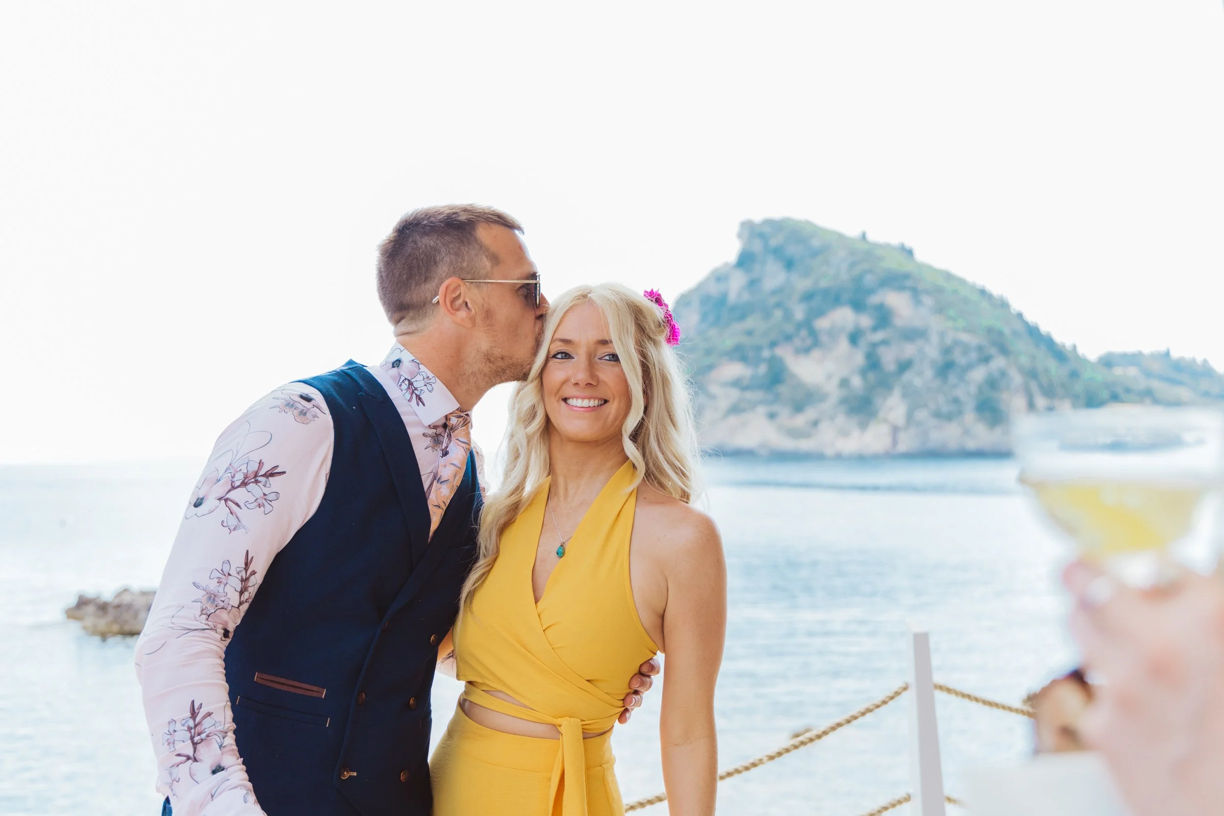 A man kisses a woman on the forehead during a celebration near the water with an island in the background, as a person holds a cocktail in the foreground.