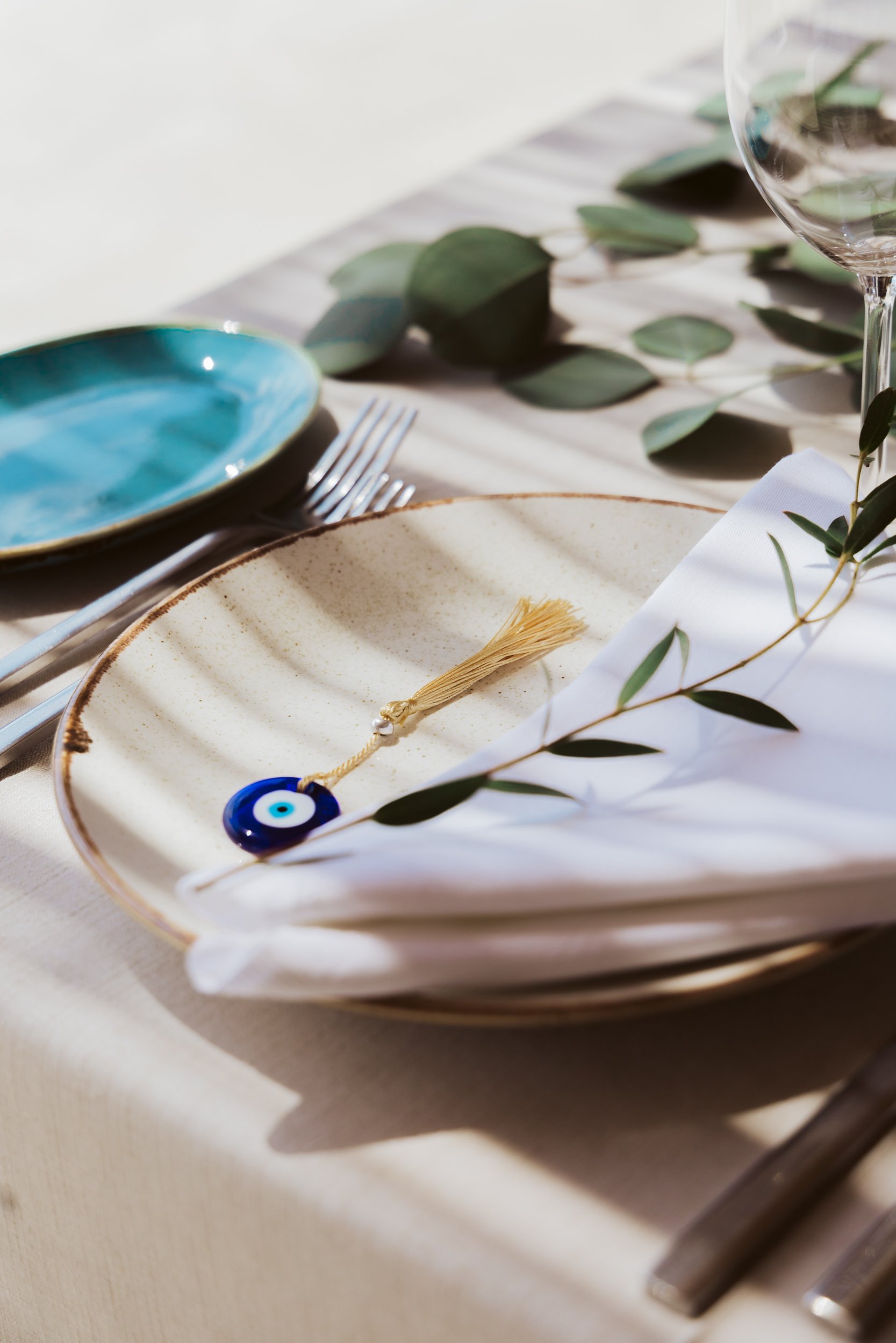 Elegant table setting with a beige ceramic plate, white napkin, green leaves, a blue glass evil eye charm, a blue plate, silverware, and a glass of water.