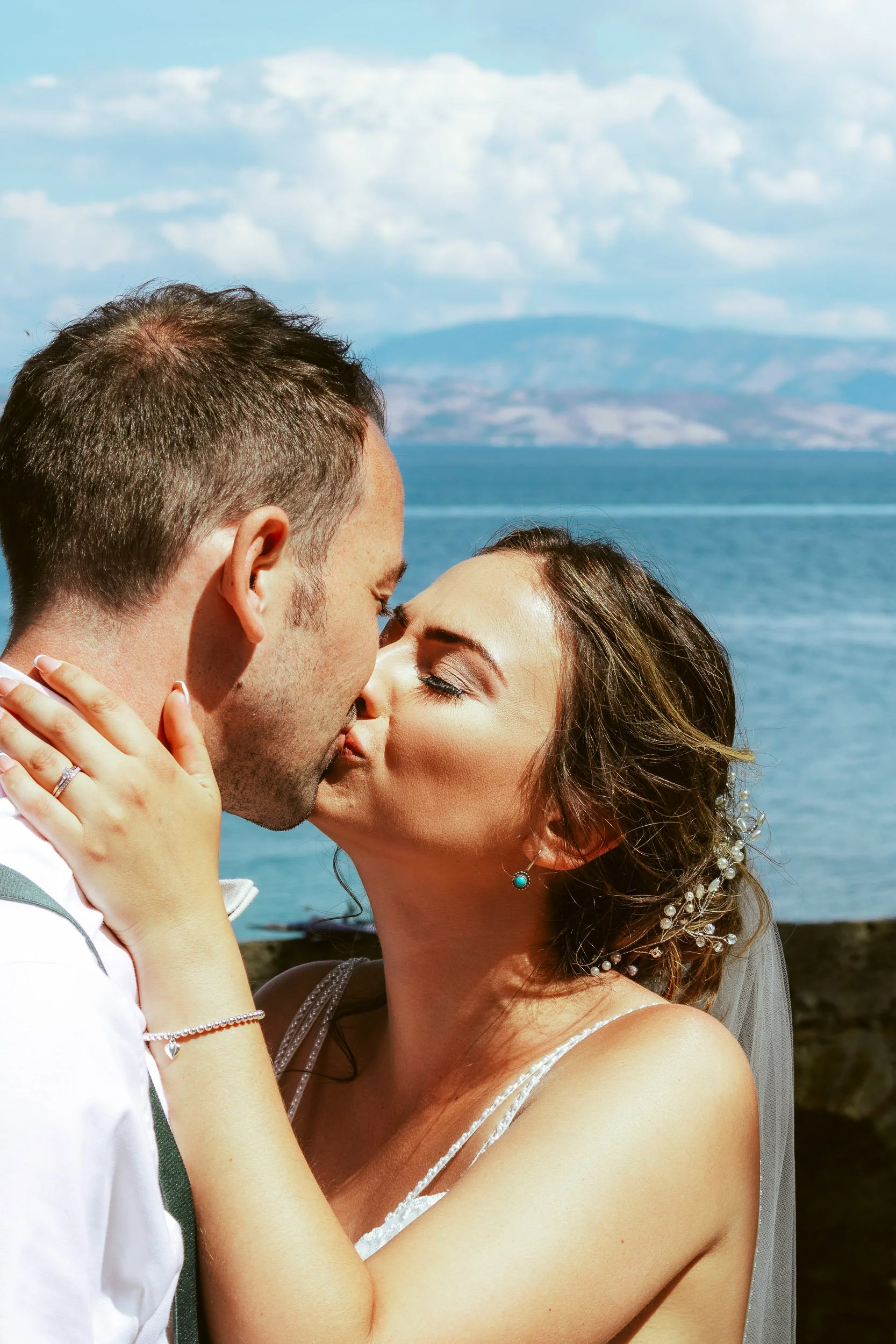 A bride and groom kissing outdoors near a body of water with mountains in the background.