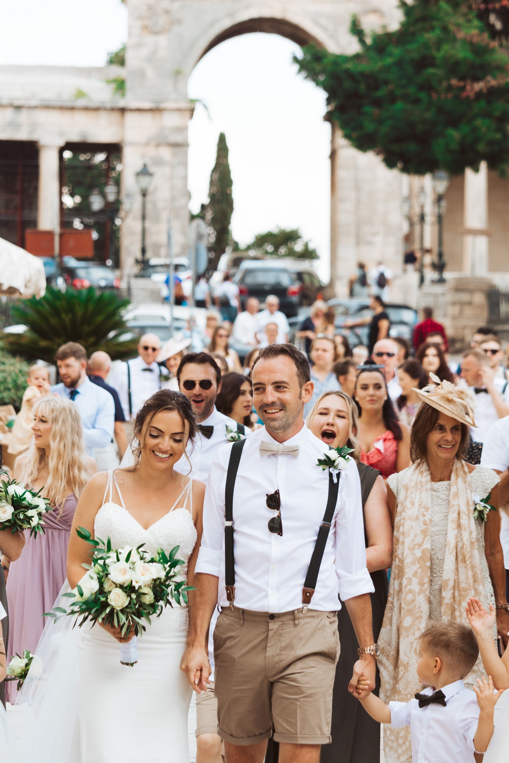 People attending a wedding procession outdoors, including a bride and groom surrounded by guests, under an ancient stone archway.