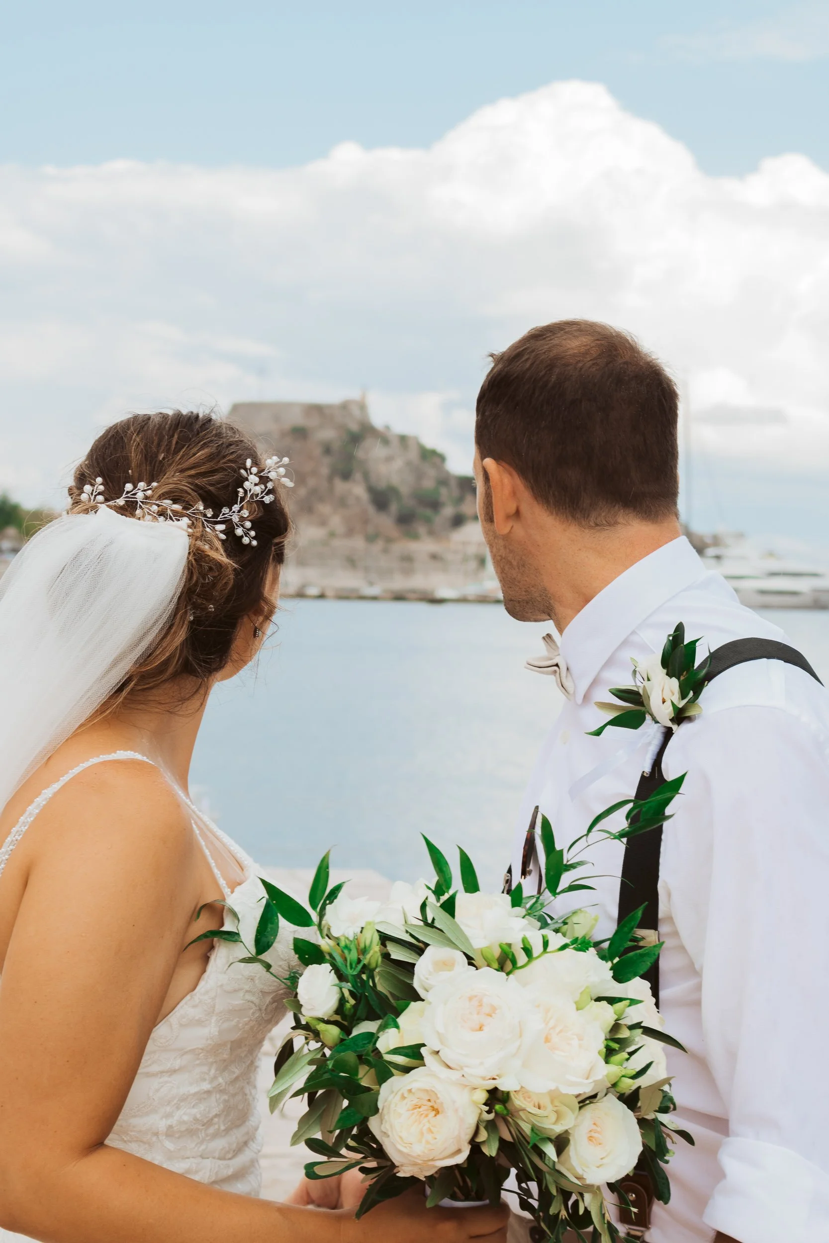 A bride and groom standing by the waterfront, looking at a castle on a hill in the distance. The bride wears a white lace wedding dress and a veil with floral headpiece, holding a large bouquet of white flowers and greenery. The groom wears a white s