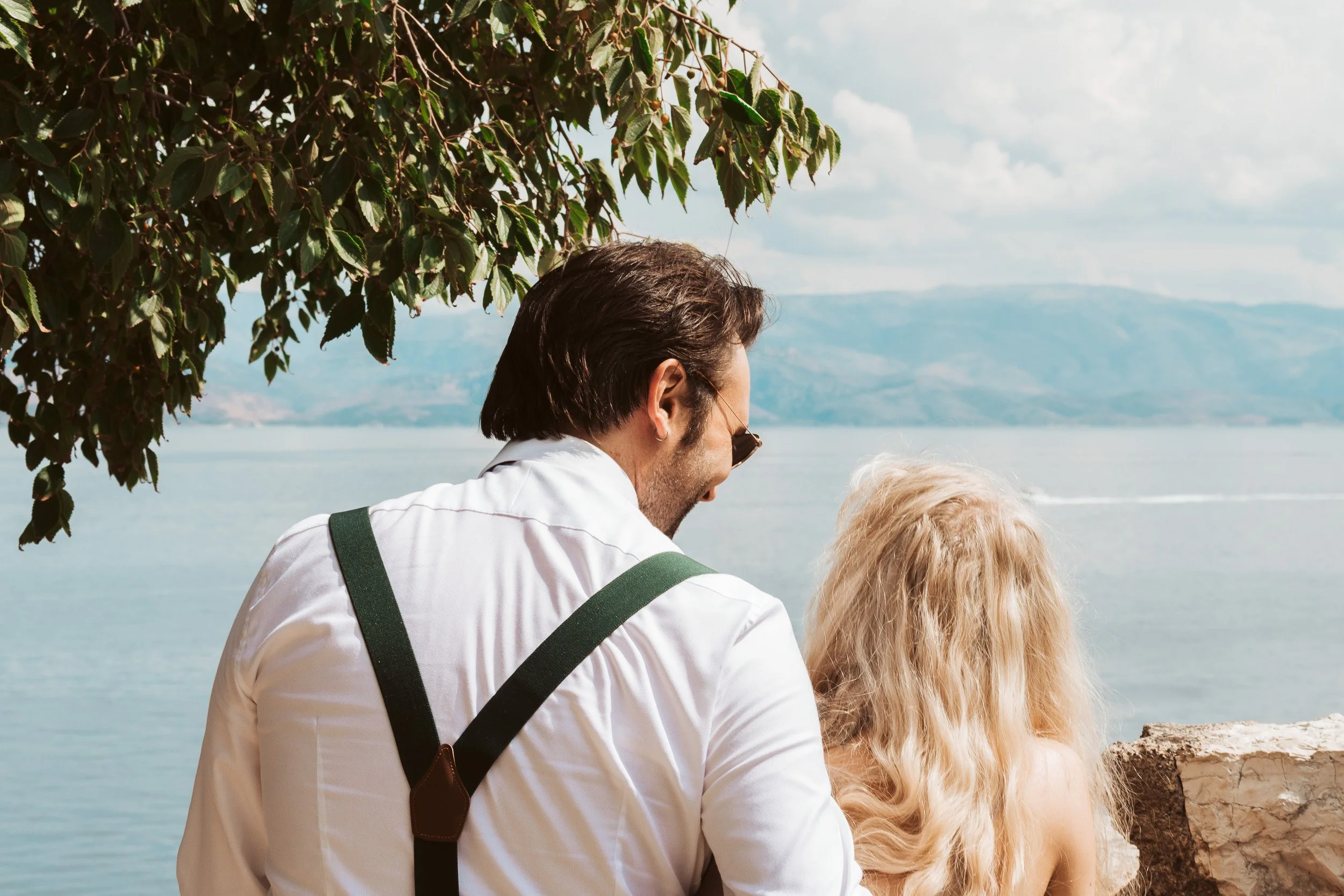 A man and a woman sitting outdoors by a lake, facing away from the camera, with mountains in the background.