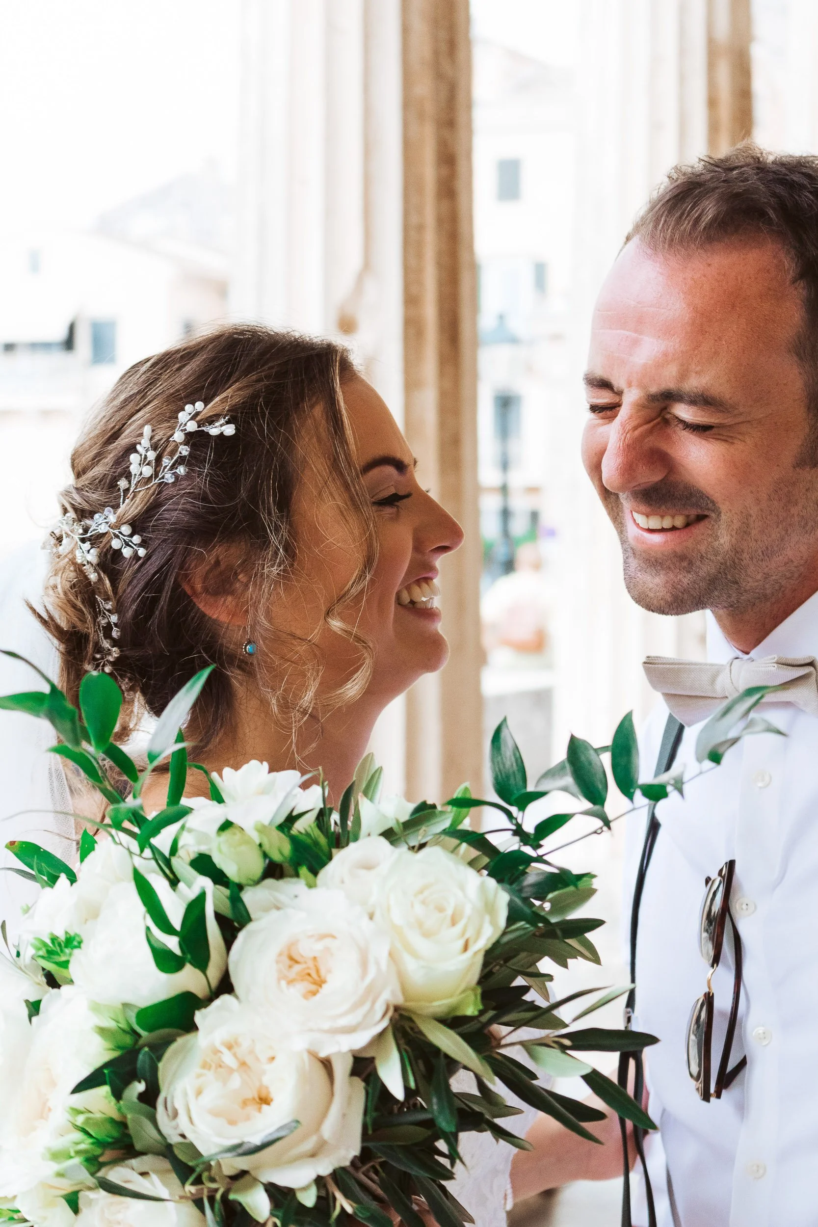A bride and groom laughing together during their wedding. The bride is holding a bouquet of white flowers and has a decorative hairpiece. The groom is wearing a white tuxedo with a bowtie.
