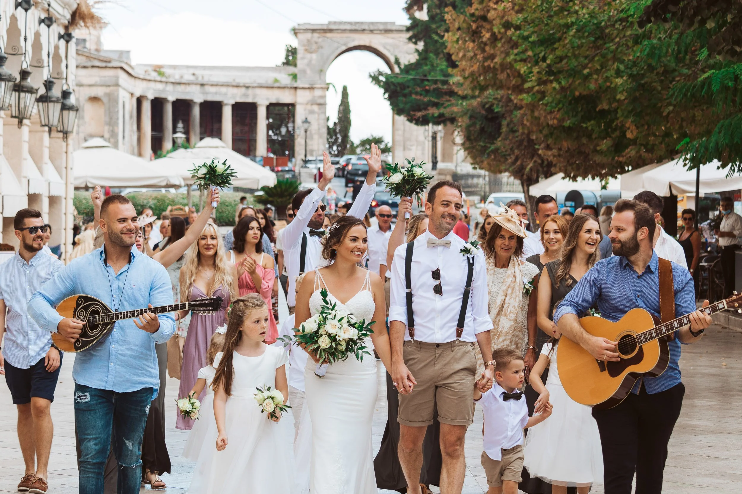 A wedding celebration with a bride and groom walking hand in hand, surrounded by friends and family, some playing guitars, outdoors in a lively setting.