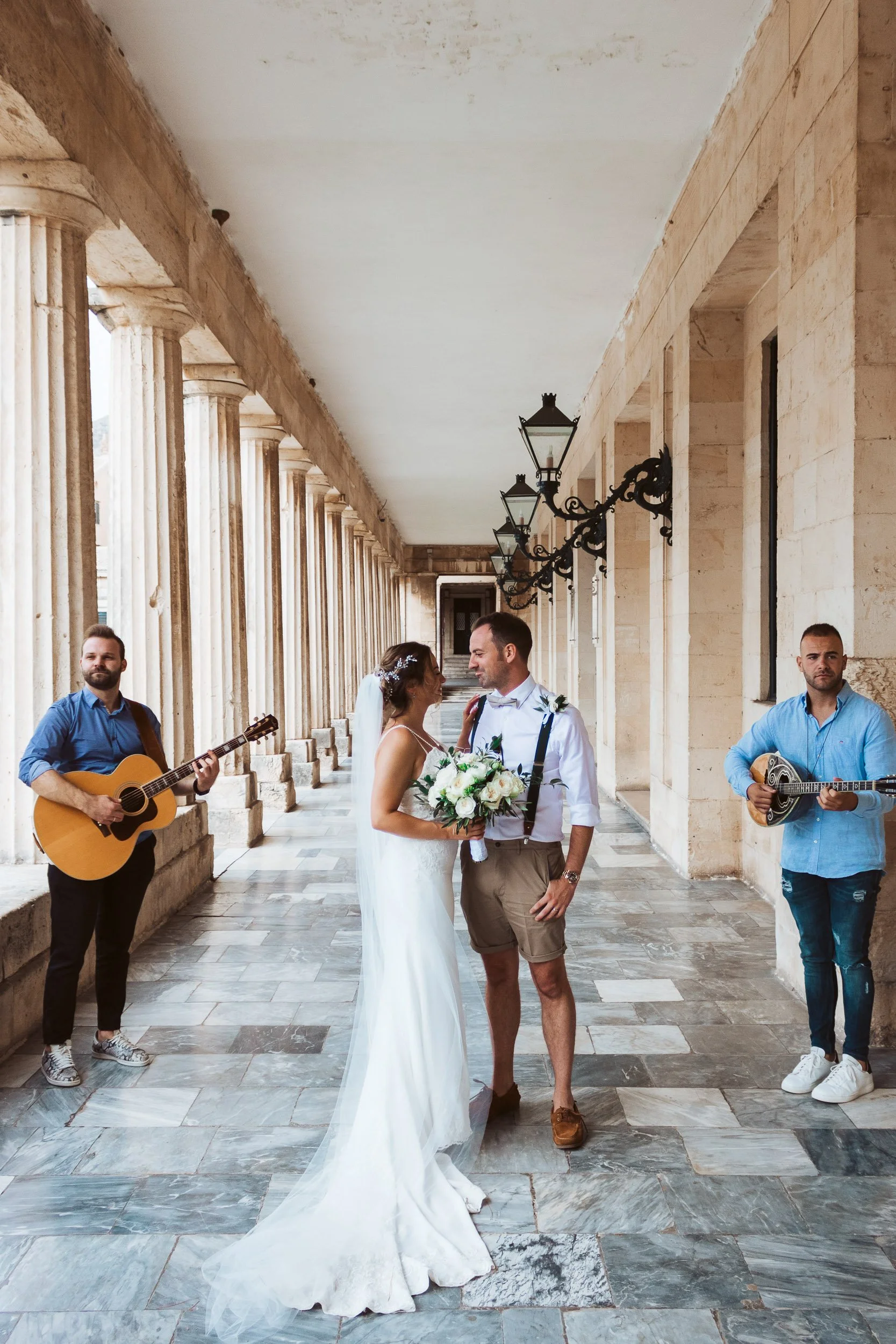 A bride and groom holding a bouquet of flowers, standing close and smiling at each other, in an outdoor corridor with stone columns and vintage black lamps. Two guitar players are on either side of them.