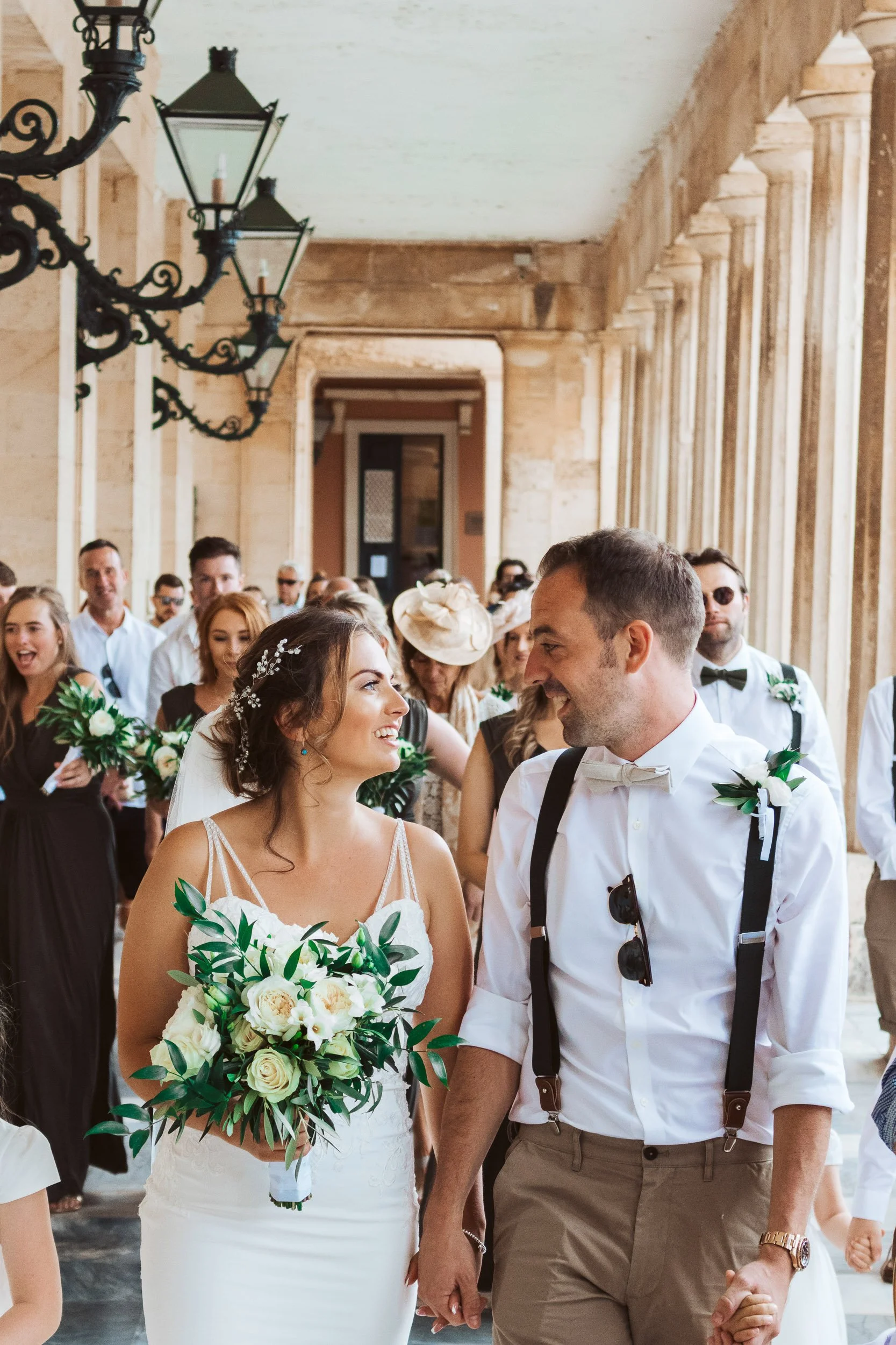 Bride and groom smiling at each other during their wedding ceremony, surrounded by guests, in an elegant hall with high ceilings, large windows, and vintage ceiling lamps.