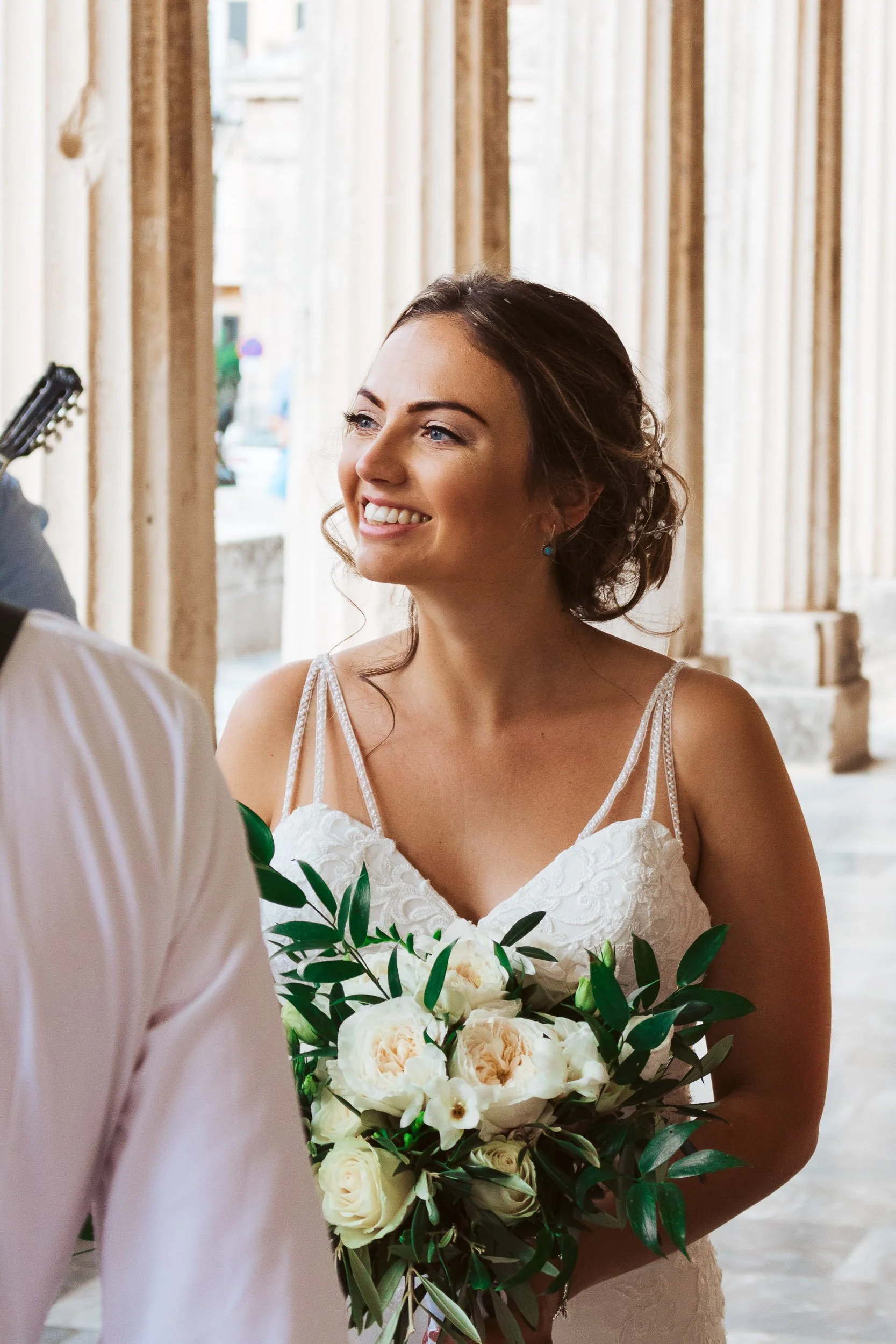 A bride holding a bouquet of white roses and greenery, smiling during her wedding ceremony indoors.