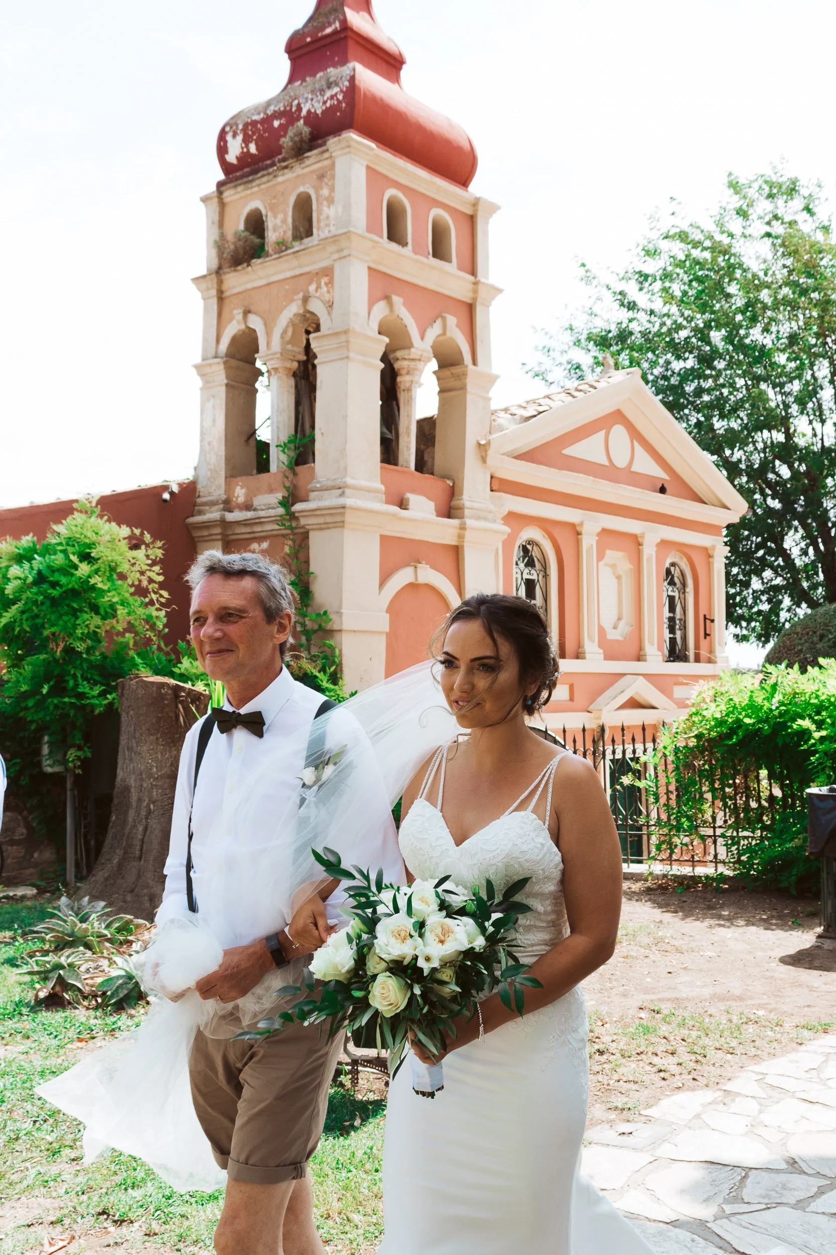 A bride holding a bouquet walking outside with an older man dressed in a white shirt and black bow tie, in front of a pink chapel with a bell tower surrounded by greenery.