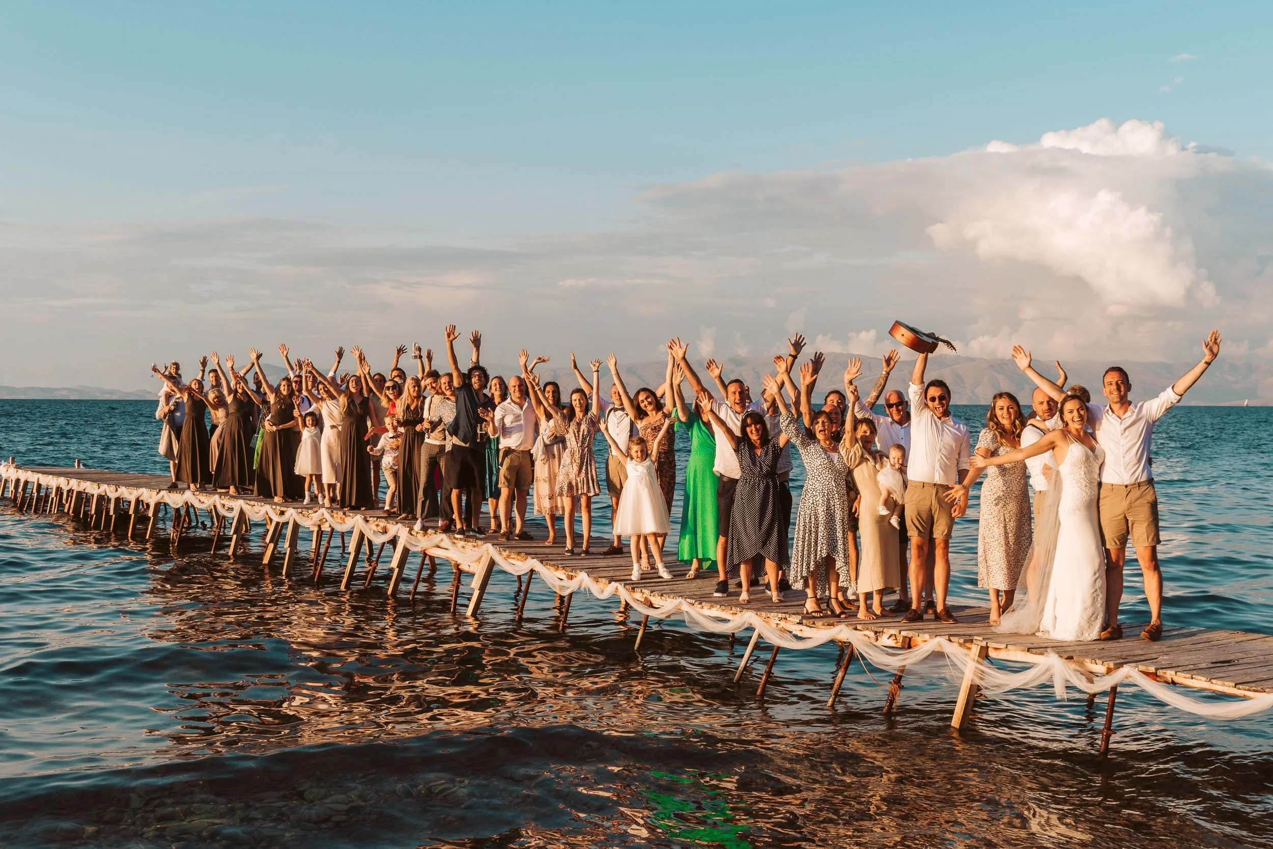 Group of people celebrating a wedding on a wooden pier extending into the water, with some women in wedding dresses, under a partly cloudy sky.