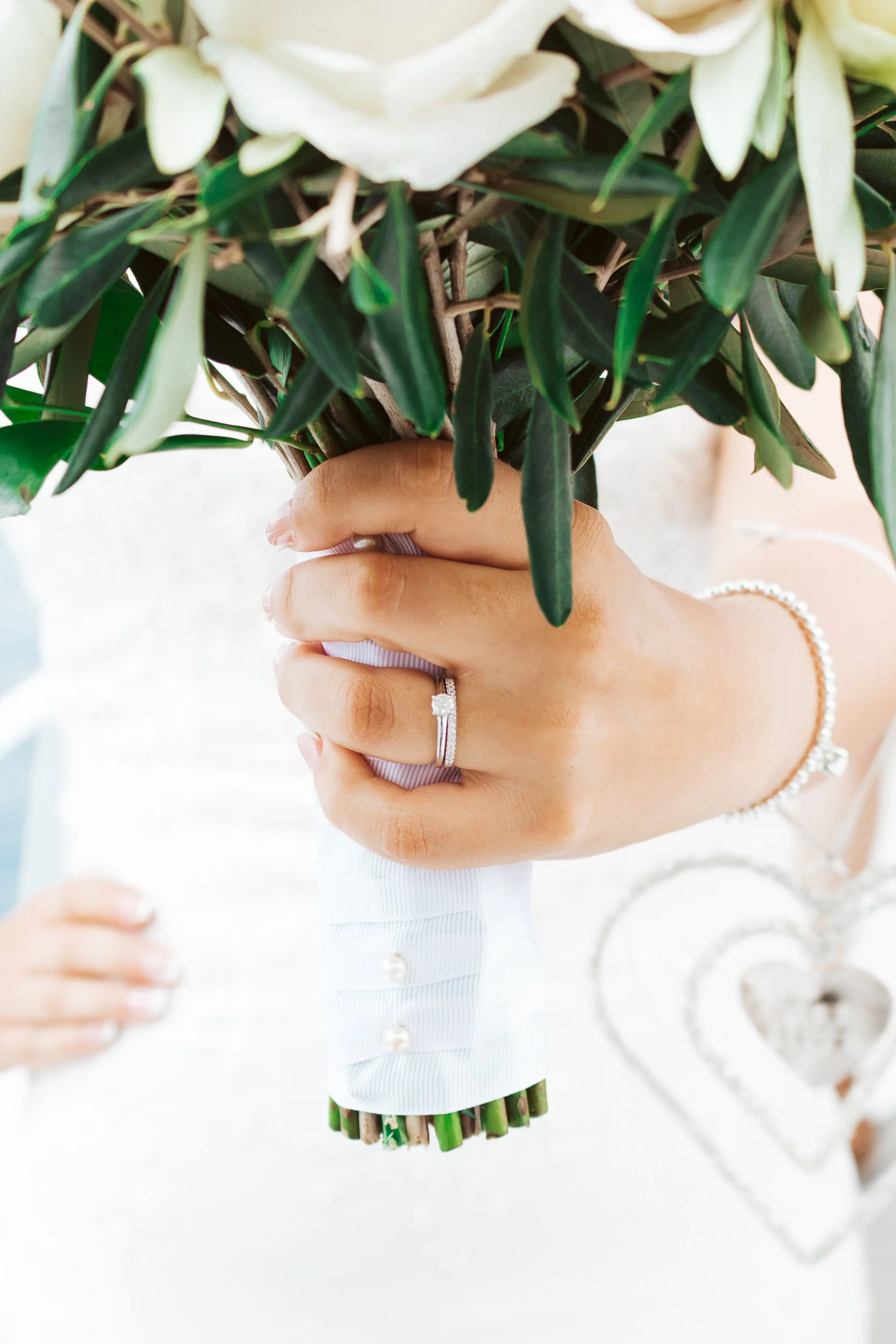 Close-up of a woman's hand holding a bouquet of white flowers and greenery, showing a wedding ring and bracelet.