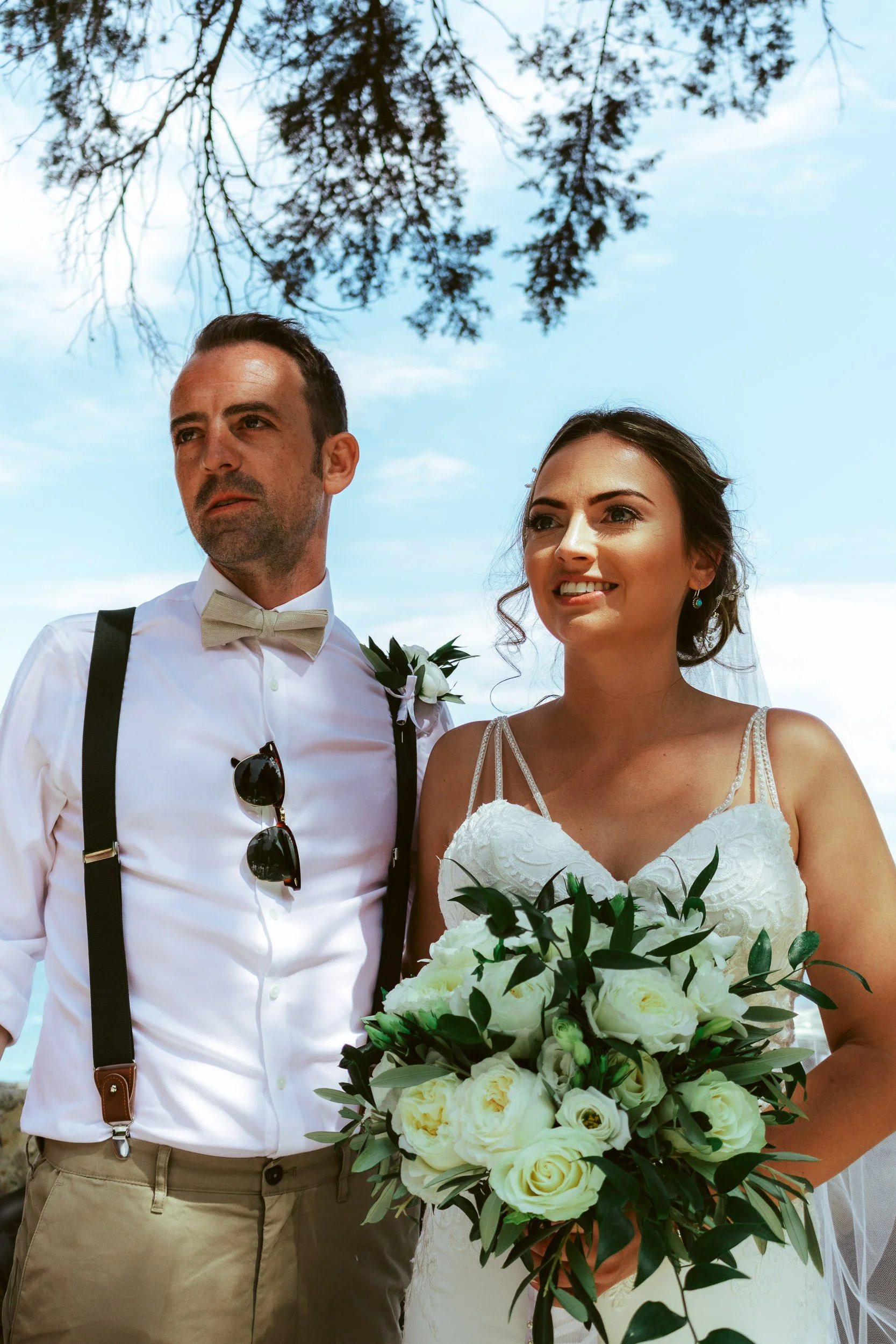 A bride and groom standing outdoors under a blue sky with tree branches overhead. The bride is holding a large bouquet of white roses and greenery, wearing a white wedding dress with thin straps. The groom is dressed in a white shirt with suspenders,