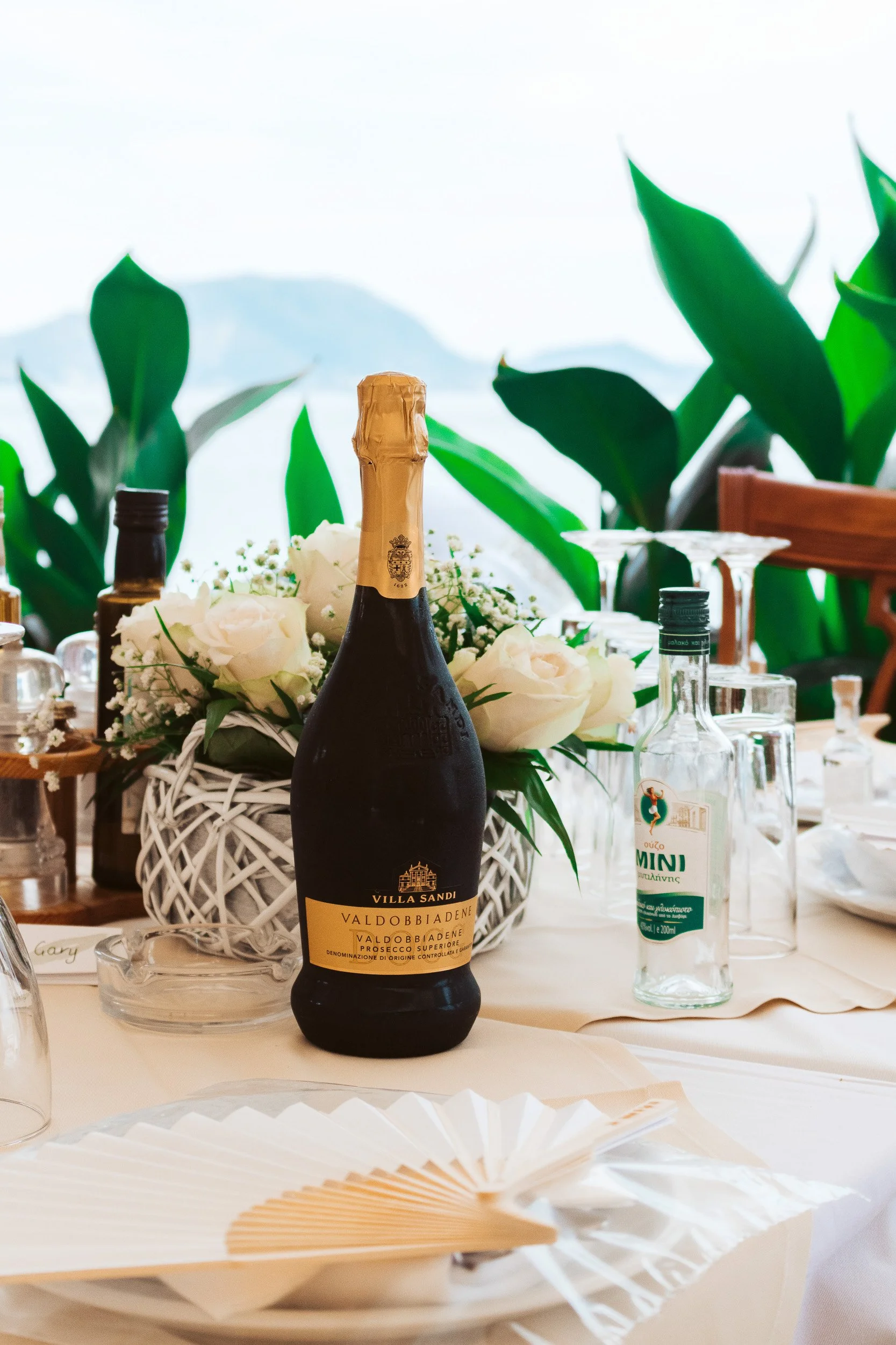 A table decorated with white flowers, a bottle of Prosecco, and various bottles of water, on an outdoor setting with green plants and mountains in the background.