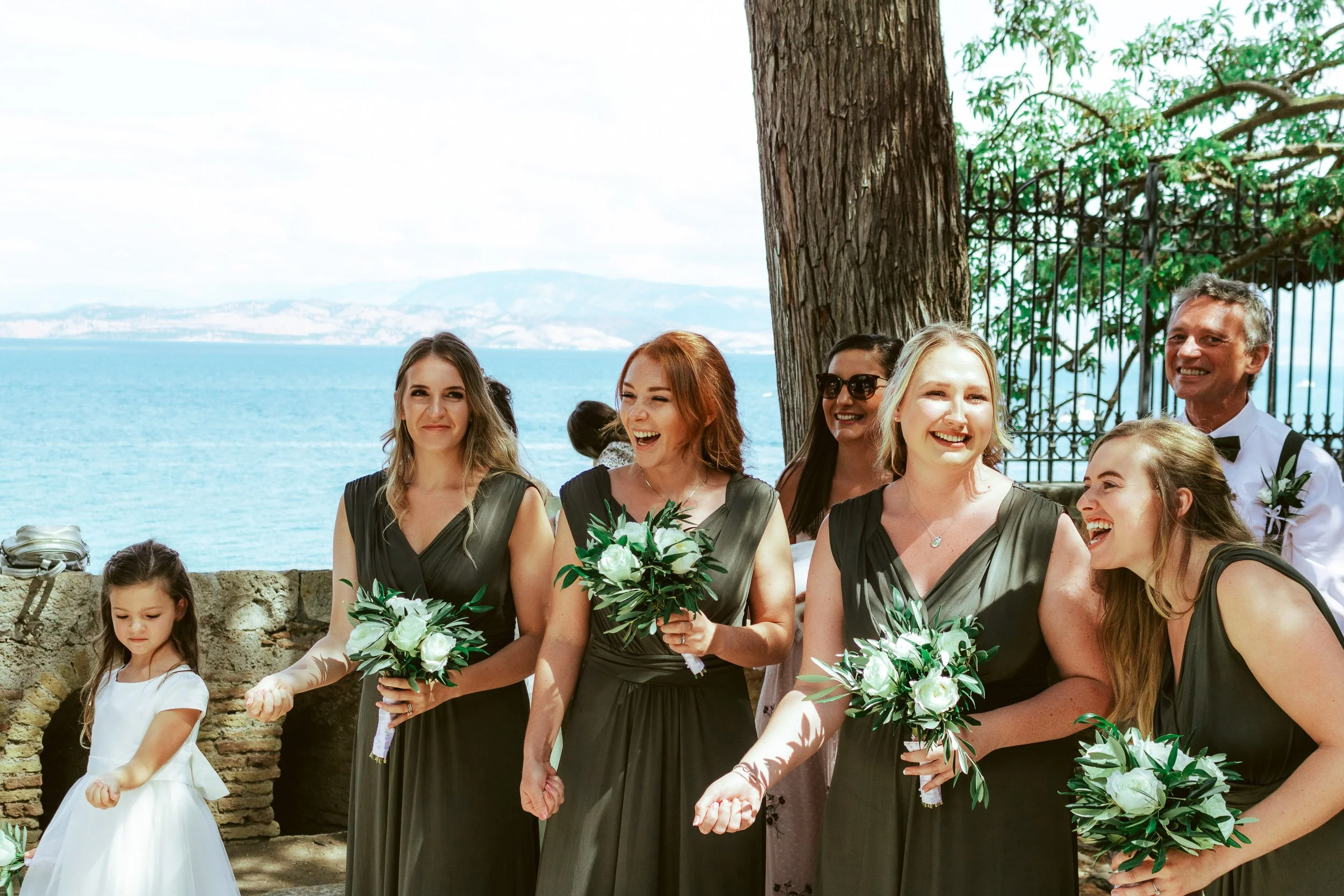 Group of women in black dresses and a young girl in white dress at a wedding ceremony outdoors by the water, holding bouquets, smiling and celebrating.