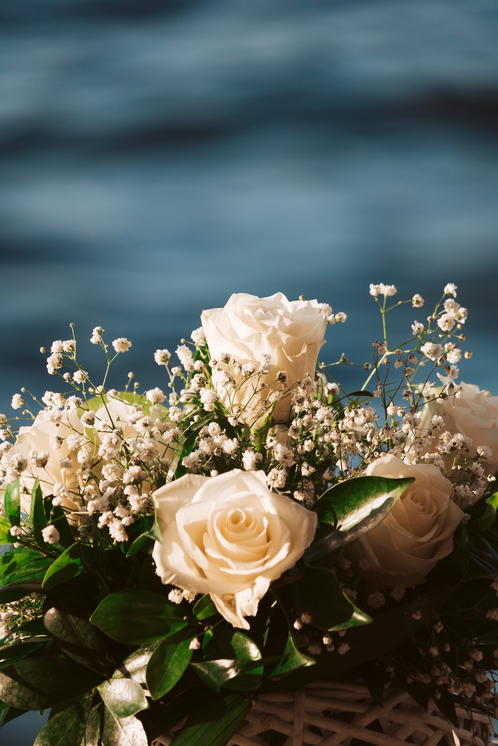 A bouquet of white roses and small white baby's breath flowers with green leaves, set against a blurred background of blue water.