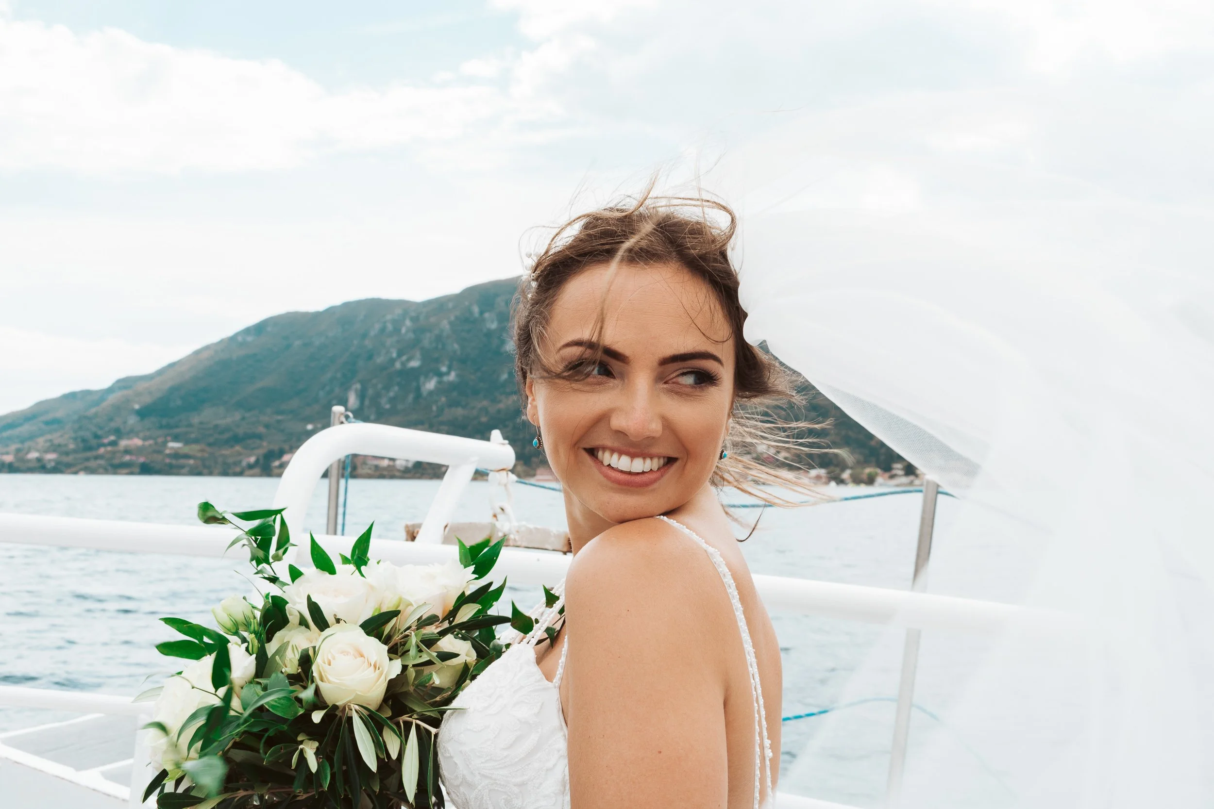 A smiling woman in a wedding dress holding a bouquet of white roses on a boat with water and mountains in the background.